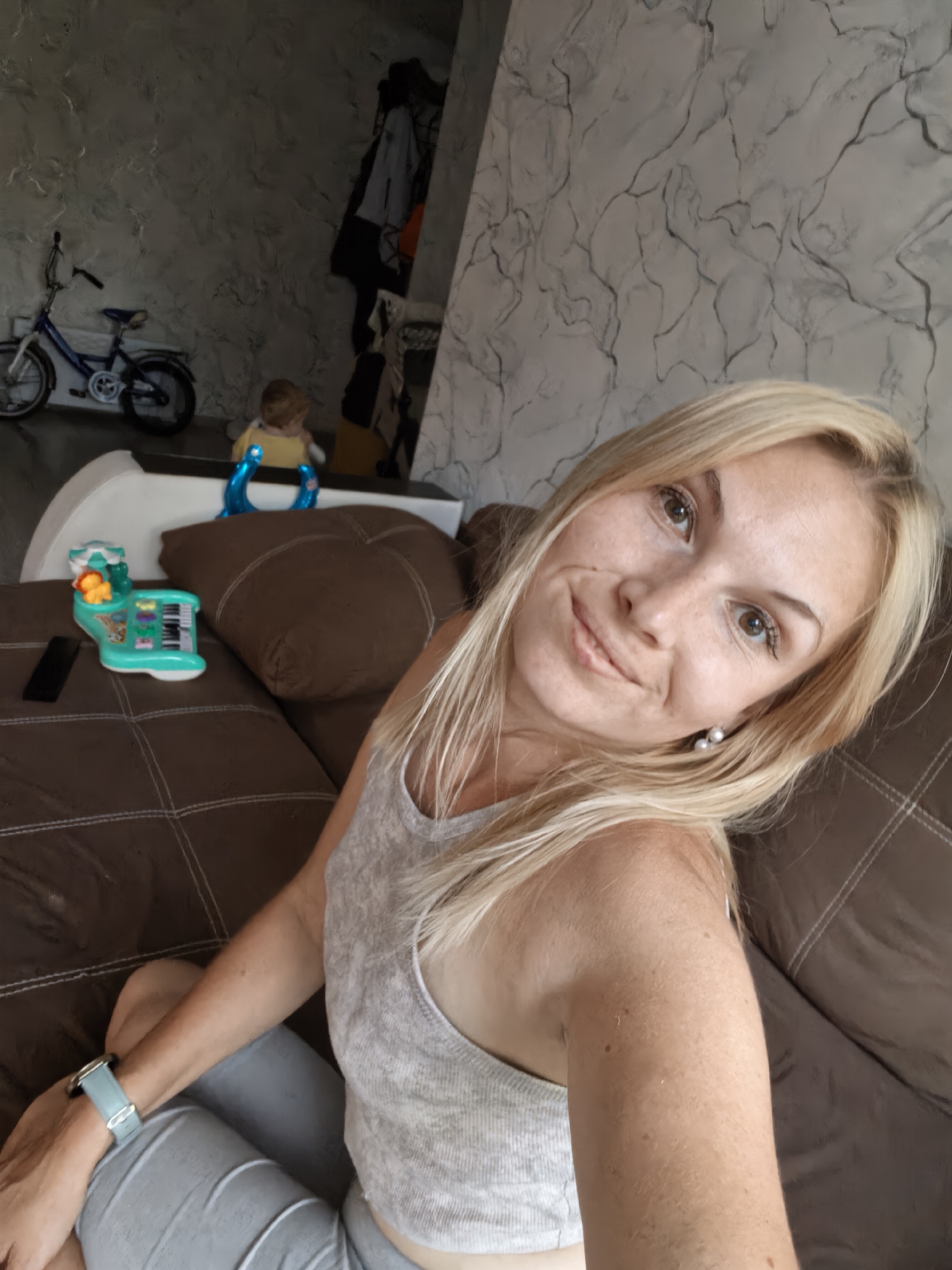 A blonde woman is taking a selfie while sitting on a brown sofa in a living room. The woman is wearing a light gray tank top and light gray pants. She has on a light blue watch. In the background, there is a small blue bicycle, a child's toy, and a child sitting in a yellow shirt. The wall in the background has a marbled pattern. The image appears to be taken indoors.