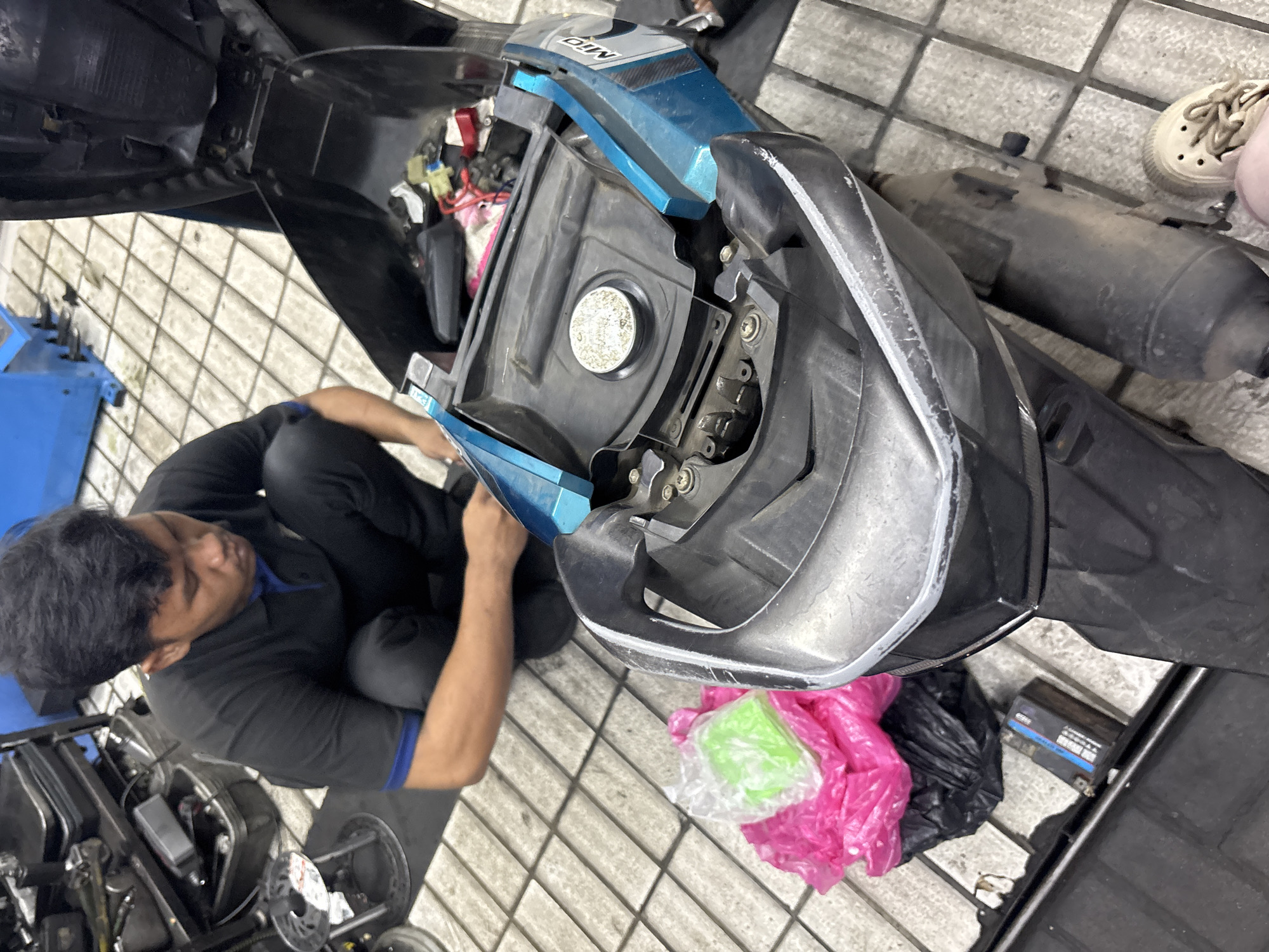 A man in a black polo shirt with a blue collar and black pants is squatting down and working on a teal and black scooter in what appears to be a workshop. The seat of the scooter is open, revealing the storage compartment underneath, which contains some wires and what look like electrical components. In the foreground, the front of the scooter is visible, including the handlebars and the area around the fuel cap. To the left of the scooter, there are two plastic bags, one pink and one black, with a small green object inside a clear plastic bag placed on top of the pink bag. The floor is tiled with a checkered pattern. In the background, a blue piece of machinery and shelves filled with tools and equipment are visible. To the right of the scooter, a person's foot wearing a beige sneaker is visible. The scene is well-lit, suggesting it is daytime. The image was taken in North Jakarta, Indonesia.