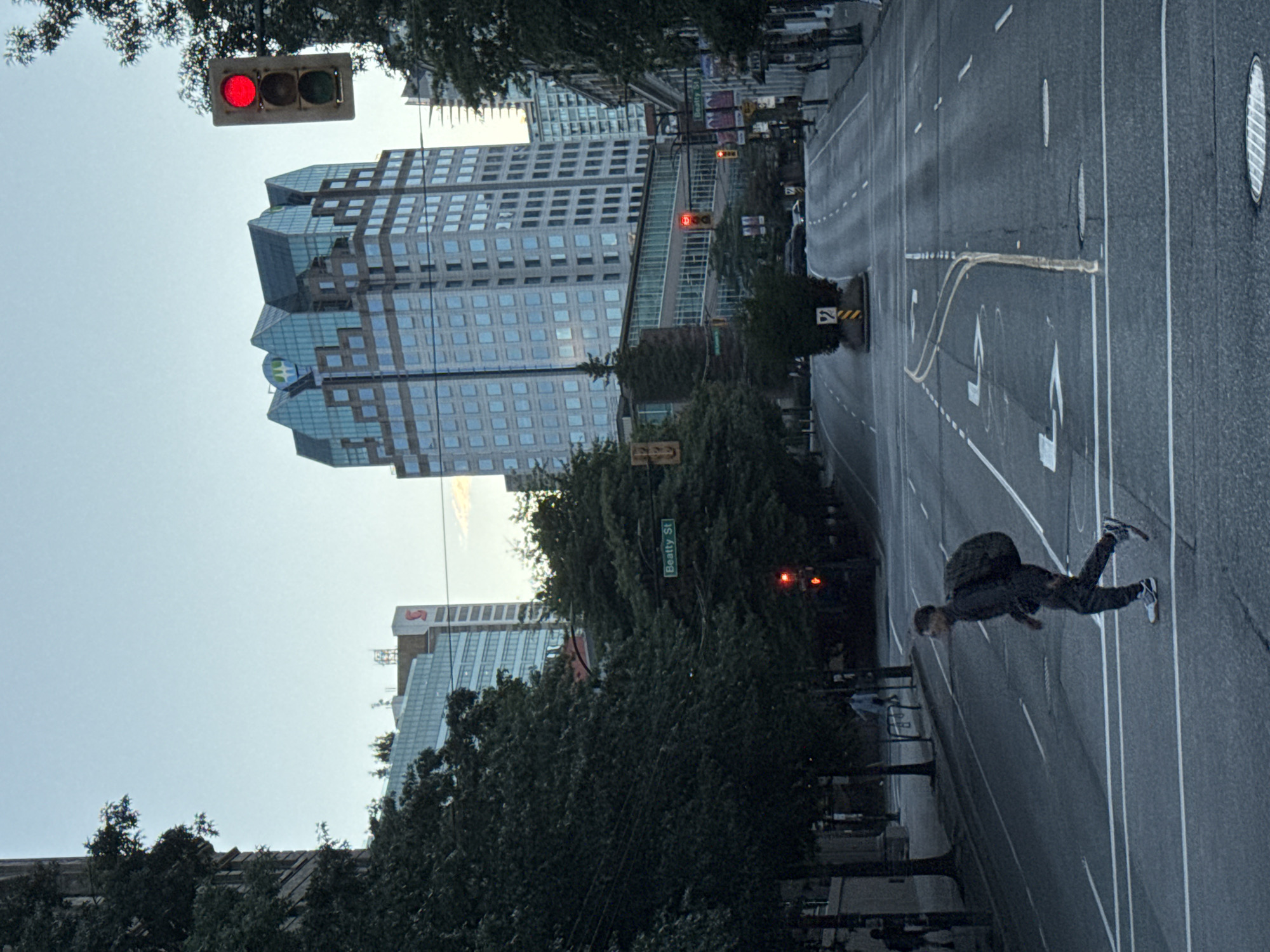 The image depicts a street scene in Vancouver, Canada. In the foreground, a young man with dark hair and a backpack is crossing a street at a pedestrian crossing. He is wearing dark clothing and white and grey sneakers. He is walking from left to right. The street is asphalt with white lane markings and a yellow dividing line. Several vehicles are visible in the distance, and one is partially obscured by a traffic island with a yellow and black striped sign.
In the middle ground, trees line the sidewalks, and street signs indicate "Beatty St." Traffic lights are visible, with one showing red. Buildings of varying heights and architectural styles are present. A notable building in the background is a tall, modern skyscraper with a distinct, multi-peaked roof structure, appearing to be glass and steel. Another building to its left is a lighter-colored structure with many windows.
The sky is clear with a pale blue hue, suggesting it is either early morning or late afternoon. The lighting is even, indicating overcast or twilight conditions. The overall impression is a typical urban streetscape in a Canadian city.