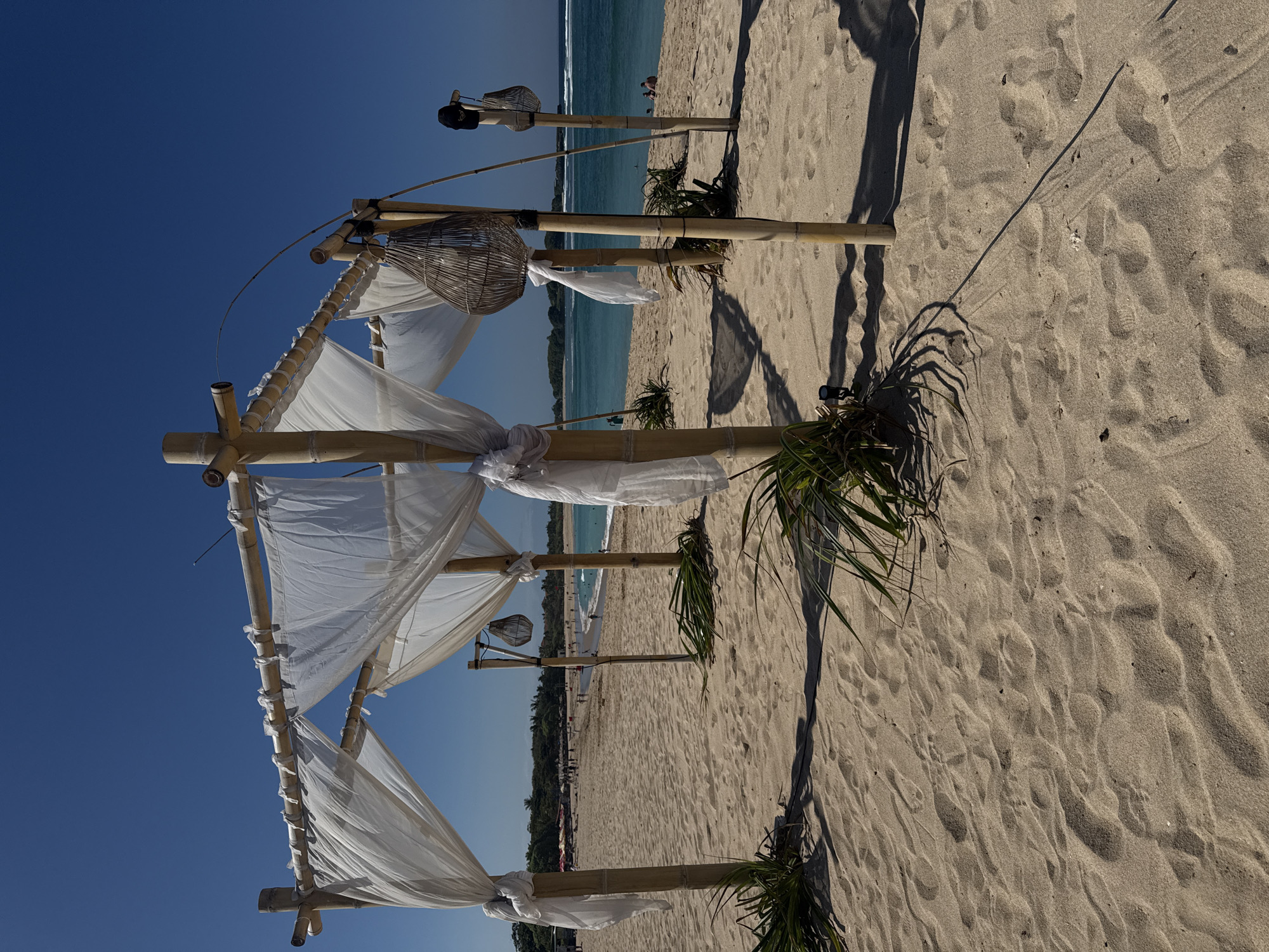 A beach scene in Kuta Selatan, Indonesia features a bamboo canopy draped with white fabric. The canopy is supported by four bamboo poles. Two woven baskets are hanging from the canopy, one of which has a black cap on it. The sand is light brown and has footprints on it. In the background, there is a turquoise ocean with gentle waves. Several people are visible in the distance on the beach and in the water. The sky is clear and blue.