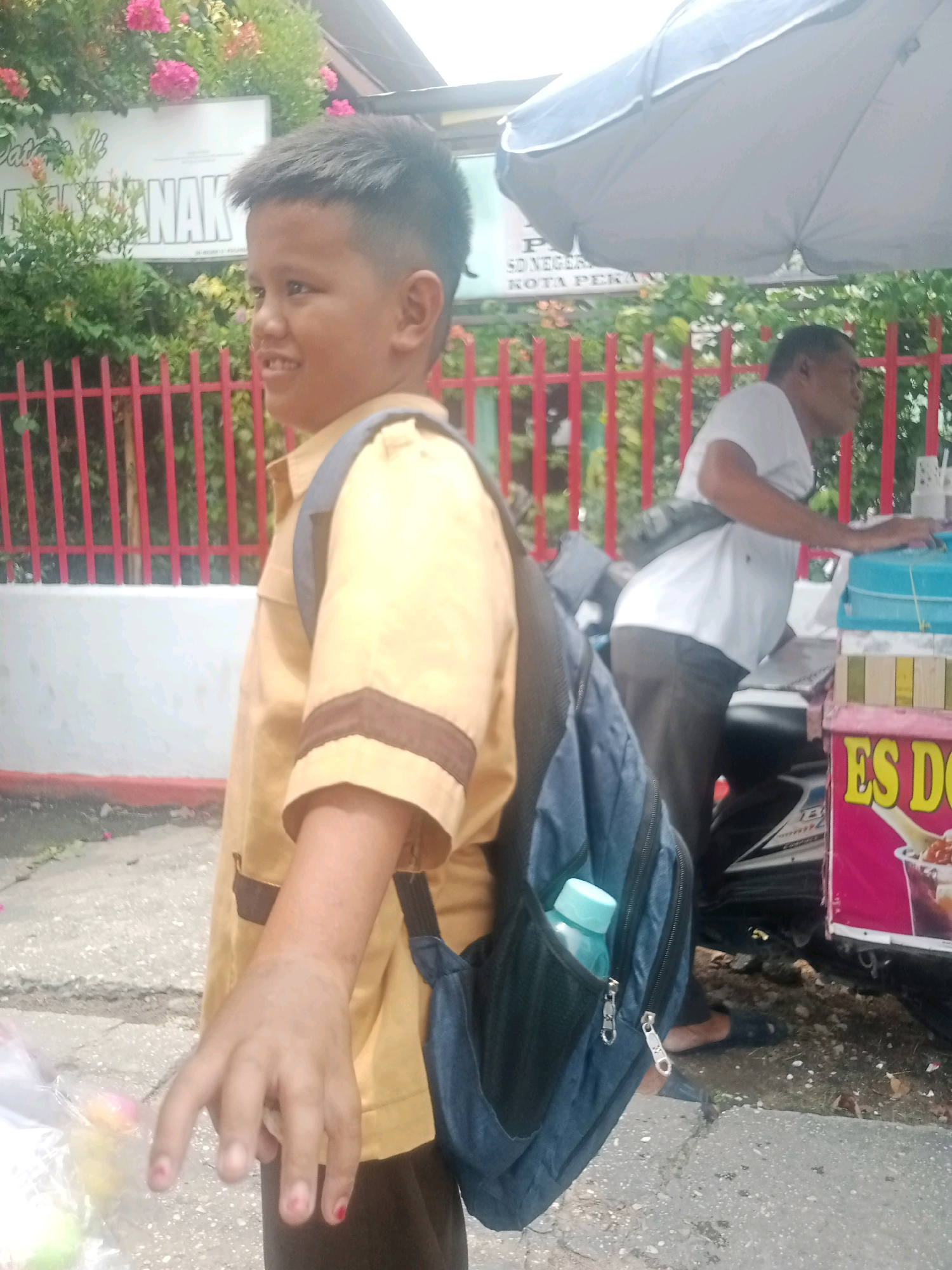 The image depicts a street scene in Pekanbaru, Indonesia. In the foreground, a young boy wearing a yellow uniform shirt and dark pants, carrying a navy blue backpack, is shown from the chest up on the left side of the frame. He is looking to his left and smiling, with his right hand extended towards the viewer. A plastic bag containing colorful objects is held by his left hand. Behind the boy, a red metal fence runs along the edge of what appears to be a school or public institution, with foliage and a sign partially visible.
In the background, an adult male in a white t-shirt and dark pants is standing next to a vendor's cart. The cart has a pink side panel with yellow text that reads "ES DO". A light blue umbrella is partially visible above the cart. The ground is paved with asphalt and concrete. The overall impression is of a casual outdoor scene likely near a school or marketplace.