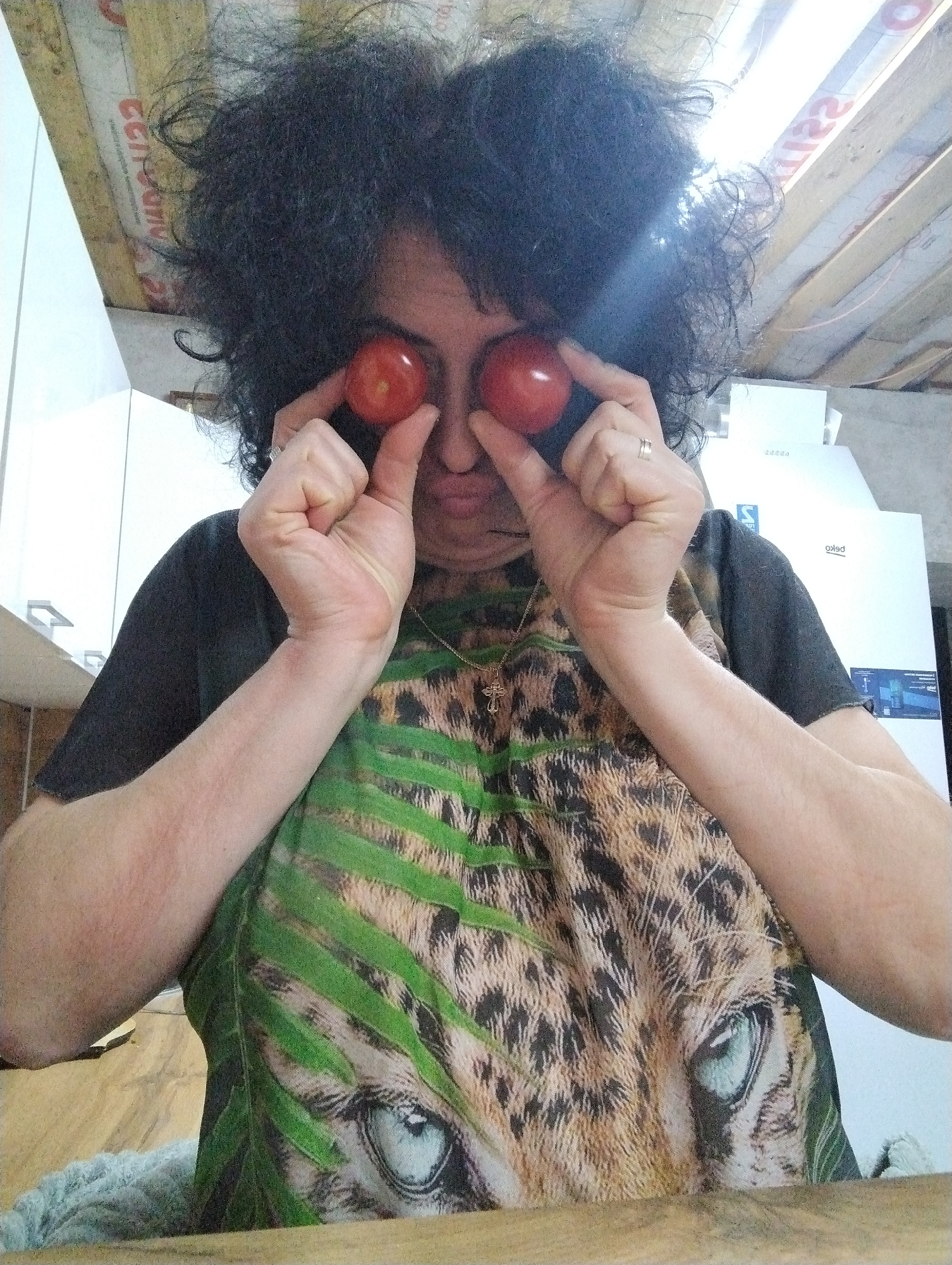 A woman with dark, curly hair is holding two red tomatoes in front of her eyes. She is wearing a black t-shirt with a print of a leopard's face surrounded by green leaves. A crucifix is visible around her neck. In the background, there is a white refrigerator and a wooden ceiling with exposed beams. The scene appears to be indoors, possibly a kitchen. There is no verifiable information to confirm the location is Grishanki, Russia.