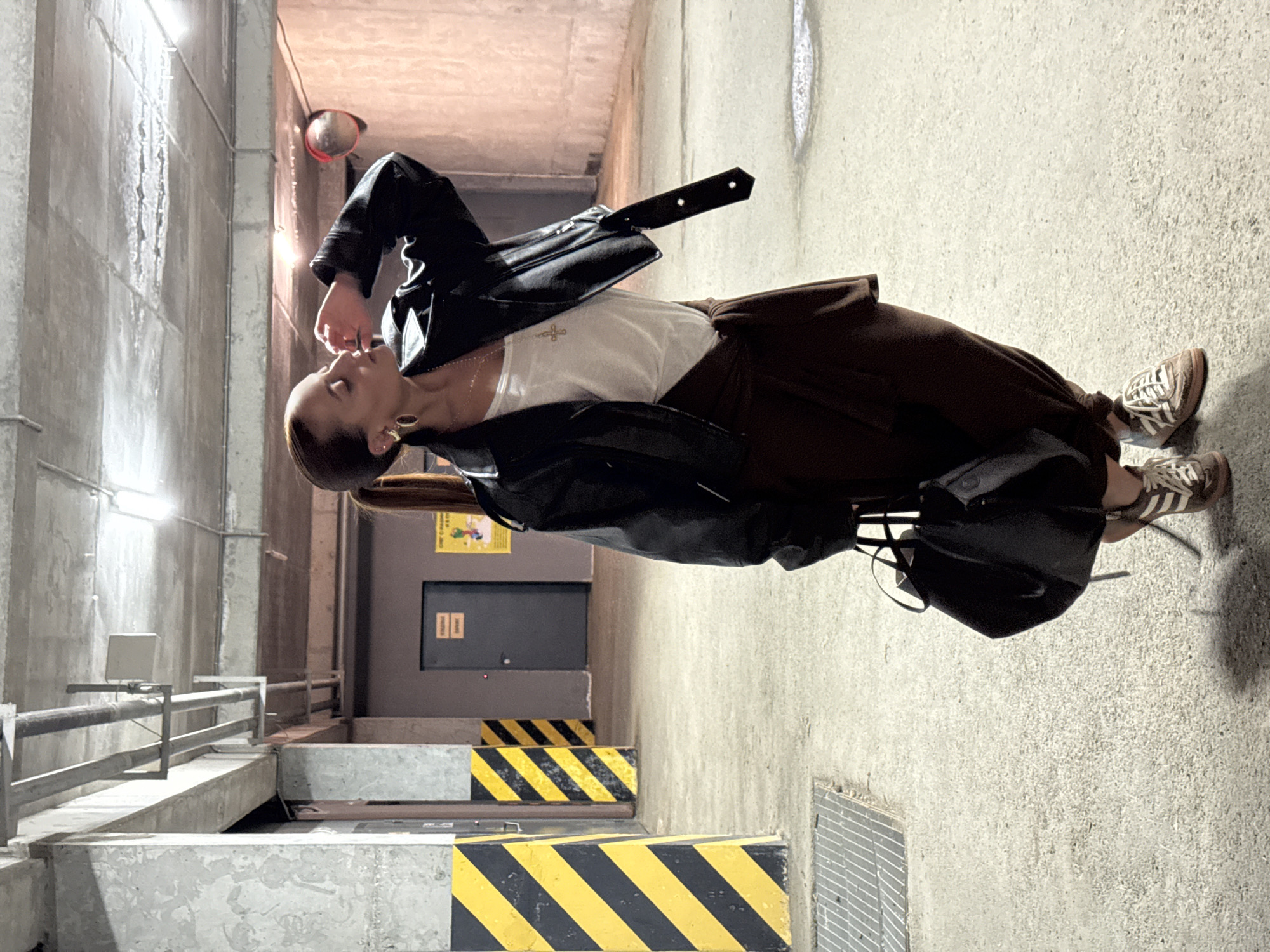A person stands in an underground parking structure. The individual wears a black leather jacket, a white top, brown pants, and Adidas sneakers. The parking structure has yellow and black striped barriers. A gray door is visible in the background. The location is Izhevsk, Russia.