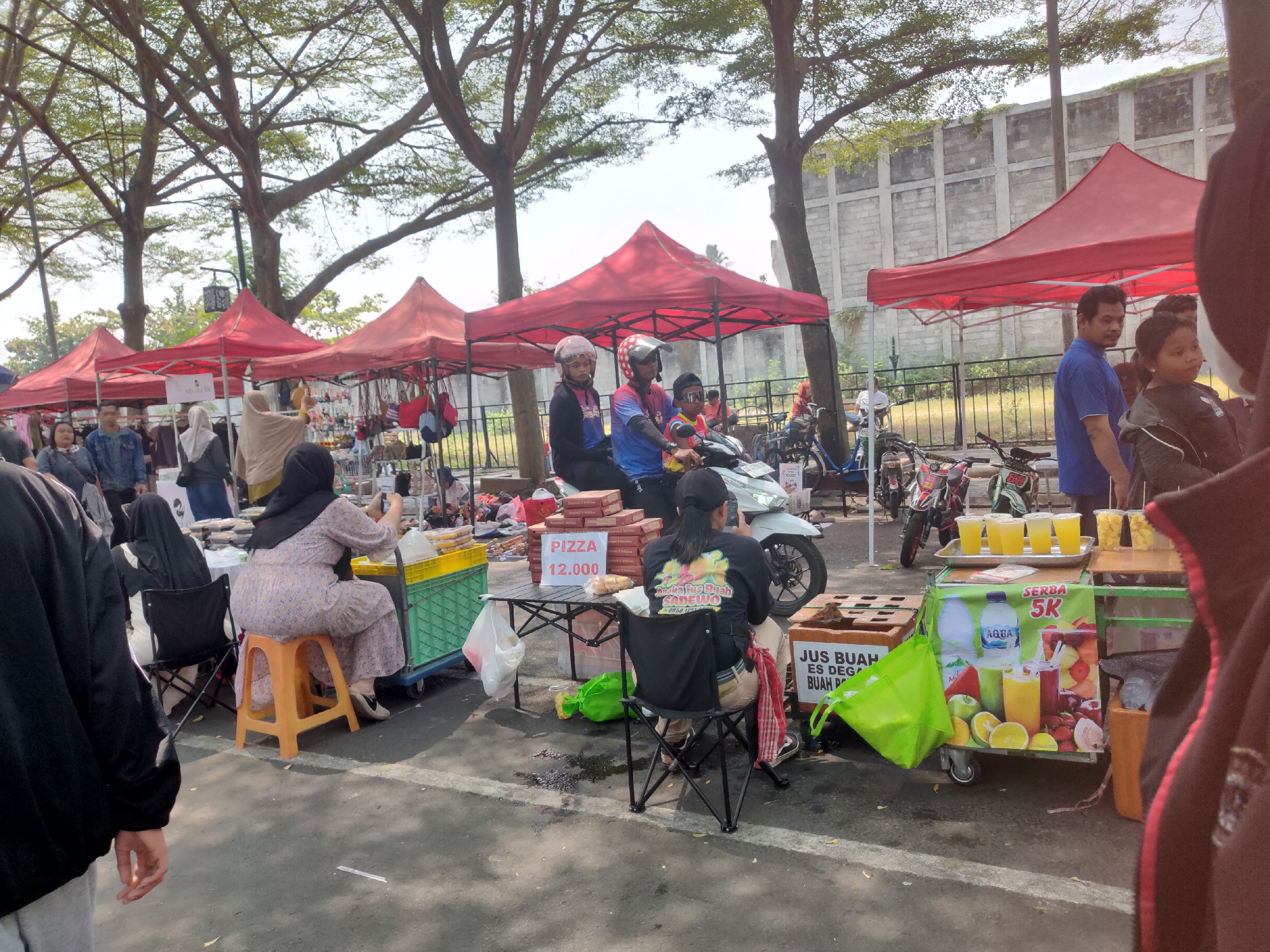 A bustling outdoor market is captured in Ngasem, Indonesia, with rows of red tents set up under the shade of large trees. Several vendors are present, offering various goods. One stall prominently displays pizzas for sale with the price of "12.000". Nearby, a juice cart is laden with colorful fruit drinks, advertising "SERBA 5K". People are seen browsing, purchasing items, and interacting with vendors. Motorcycles are parked in the background, suggesting a busy day for the market. The overall atmosphere appears lively and active, typical of a local Indonesian marketplace.