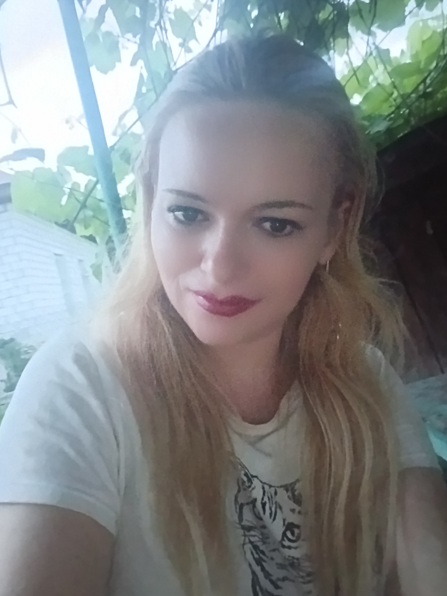 A woman with blonde hair and dark eye makeup is pictured from the chest up. She is wearing a white t-shirt with a tiger graphic. She is outdoors, with green leaves and branches overhead. Behind her to the left is a white brick wall and a blue pole. The lighting is bright and appears to be natural daylight. The image is slightly blurry and taken from a low angle.