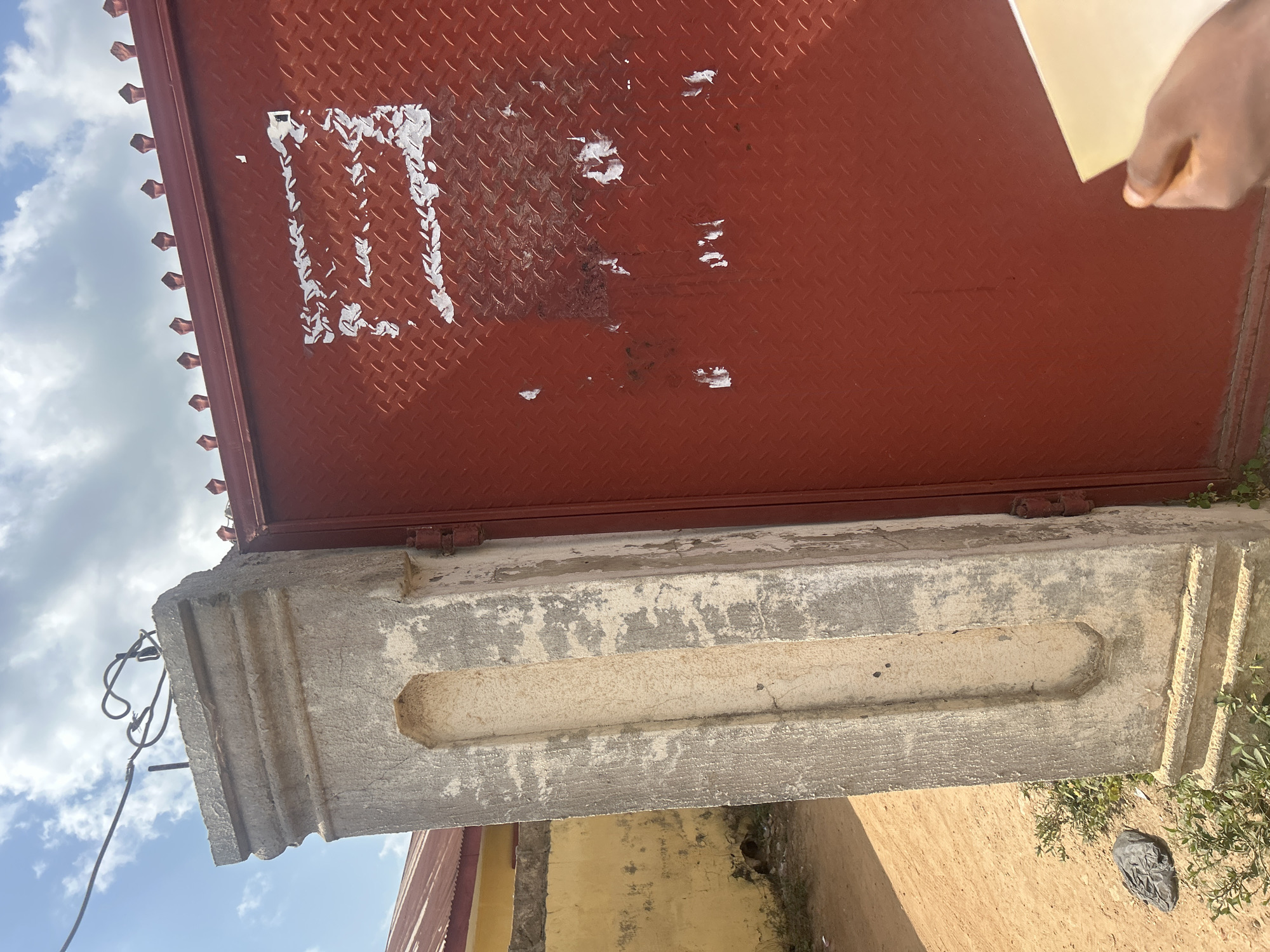 The image shows a section of a red diamond plate metal gate with some torn posters on it, attached to a grey concrete pillar. The pillar has a decorative recessed section and is weathered. Behind the pillar, a yellow wall is visible with a red-roofed building. The ground is sandy with some green vegetation and a black plastic bag. The sky is partly cloudy. A person's hand is visible in the bottom right corner, holding a light-colored rectangular object. This scene is located in Ogbomosho, Nigeria.