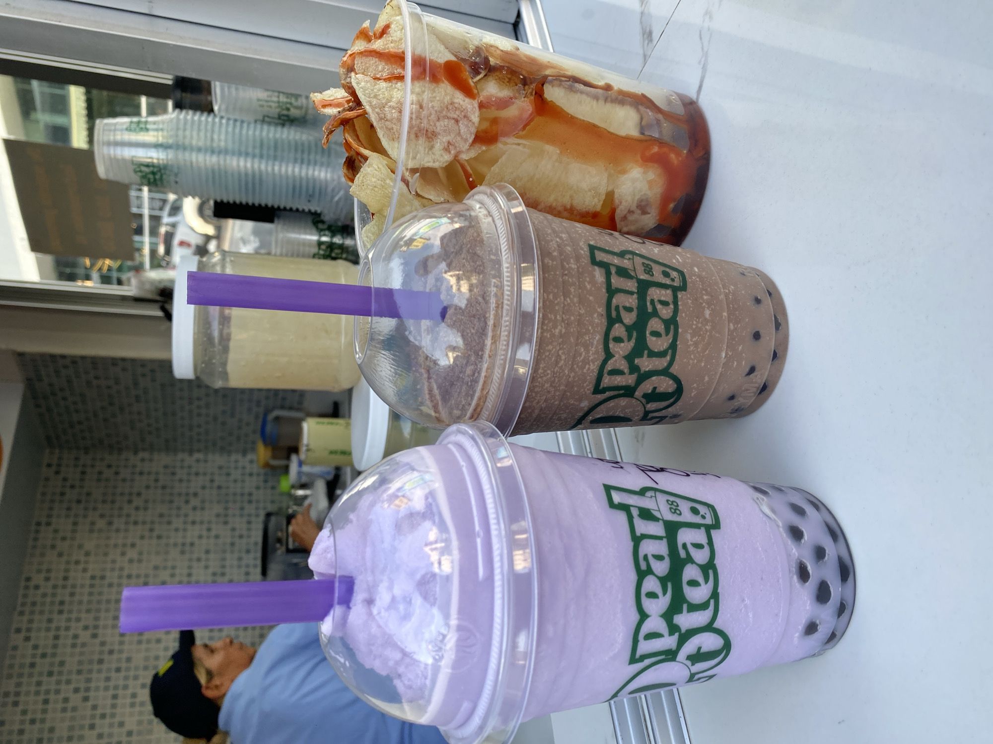 The image captures a close-up of three beverages, likely boba teas, arranged on a white surface. The beverages are in clear plastic cups with lids and straws. The first beverage at the bottom appears to be a purple-colored boba tea with black tapioca pearls. The second beverage is a chocolate boba tea. The top beverage is a mixture of potato chips and sauce. The beverages have logos printed on them. In the background, a person wearing a blue shirt and a baseball cap is visible, and the background appears to be a food service area with assorted containers and equipment. The beverages are likely at a Pearl Tea 88 location, but there is no clear identifier of the city.