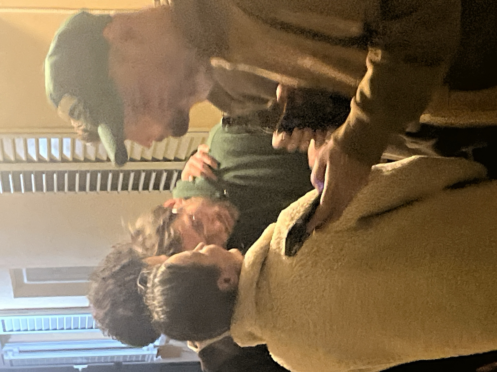 The image depicts four individuals in an indoor setting. One person, wearing a green cap and a brown jacket, is bent over, interacting with the other three. One of the three is wearing a cream-colored jacket, and another is wearing a green shirt. The location is unspecified and there is no indication that this image was taken in Tivat, Montenegro.