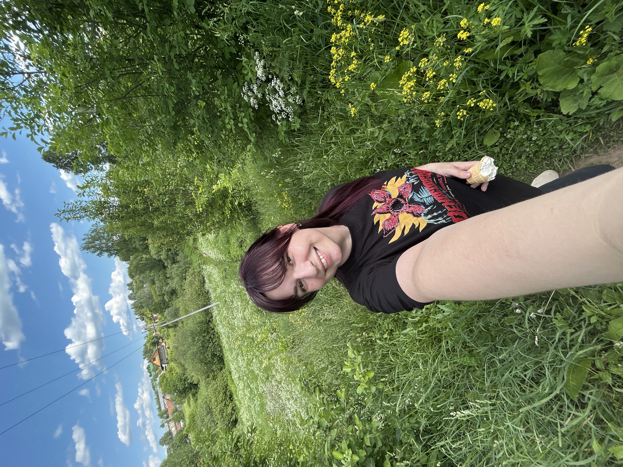 This is a portrait-style, selfie photograph taken outdoors in a grassy, rural setting. The primary subject is a young woman with dark, shoulder-length hair and bangs, smiling at the camera. She is wearing a black t-shirt with a colorful graphic print featuring a stylized creature. In her right hand, she holds a partially eaten ice cream cone in a waffle cup.
The background is dominated by lush green foliage, including tall grasses and trees. Yellow and white wildflowers are scattered throughout the grass, adding pops of color to the scene. In the distance, beyond a line of trees, hints of houses and a power line are visible, suggesting a village or small town. The sky is a vibrant blue with scattered white, fluffy clouds.
The lighting suggests it is daytime, likely late morning or afternoon, under sunny weather conditions. The overall mood is cheerful and relaxed, with the woman appearing happy and enjoying a treat outdoors. The photograph is oriented vertically. The image contains no visible text. The location context provided is Фоки, Russia.