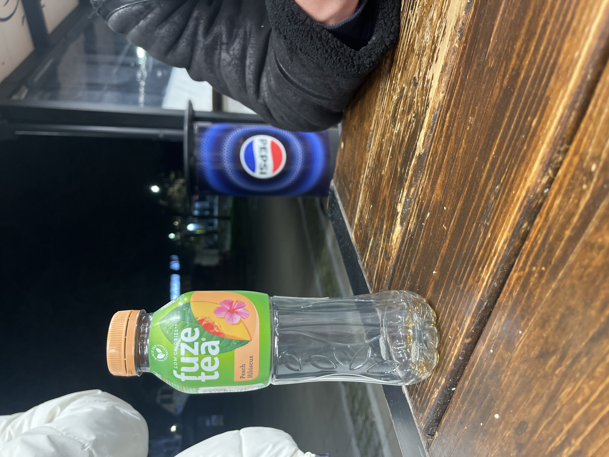 Here is a description of the image:
The image shows a partially consumed bottle of Fuze Tea, Peach Hibiscus flavor, resting on a wooden surface with a visible grain pattern. To the left of the bottle, there is a Pepsi-branded trash can, suggesting an outdoor or semi-outdoor setting. A person wearing a dark, possibly leather or fur-lined jacket is partially visible in the upper portion of the image. In the lower left, a section of a white garment is also visible. The background features a night-time cityscape with streetlights and possibly some buildings, although details are obscured by the darkness. Overall, the scene appears to be a casual, perhaps temporary outdoor dining or seating area.