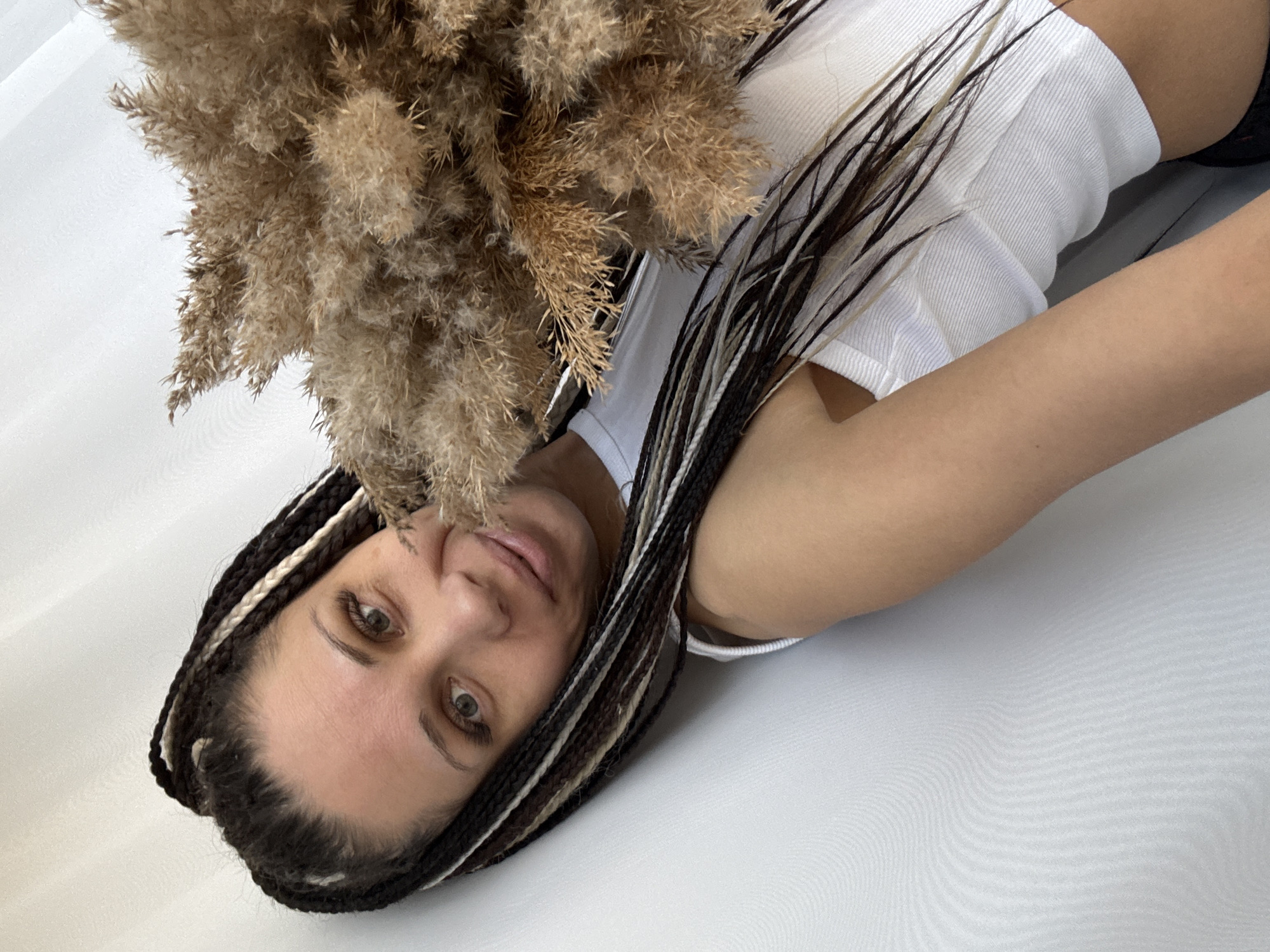 A woman with black and white braids is holding a bouquet of dried flowers. She is wearing a white tank top. The background appears to be a white surface, possibly a sheet or wall. The image's orientation is rotated. The location is Rusanovo, Russia.