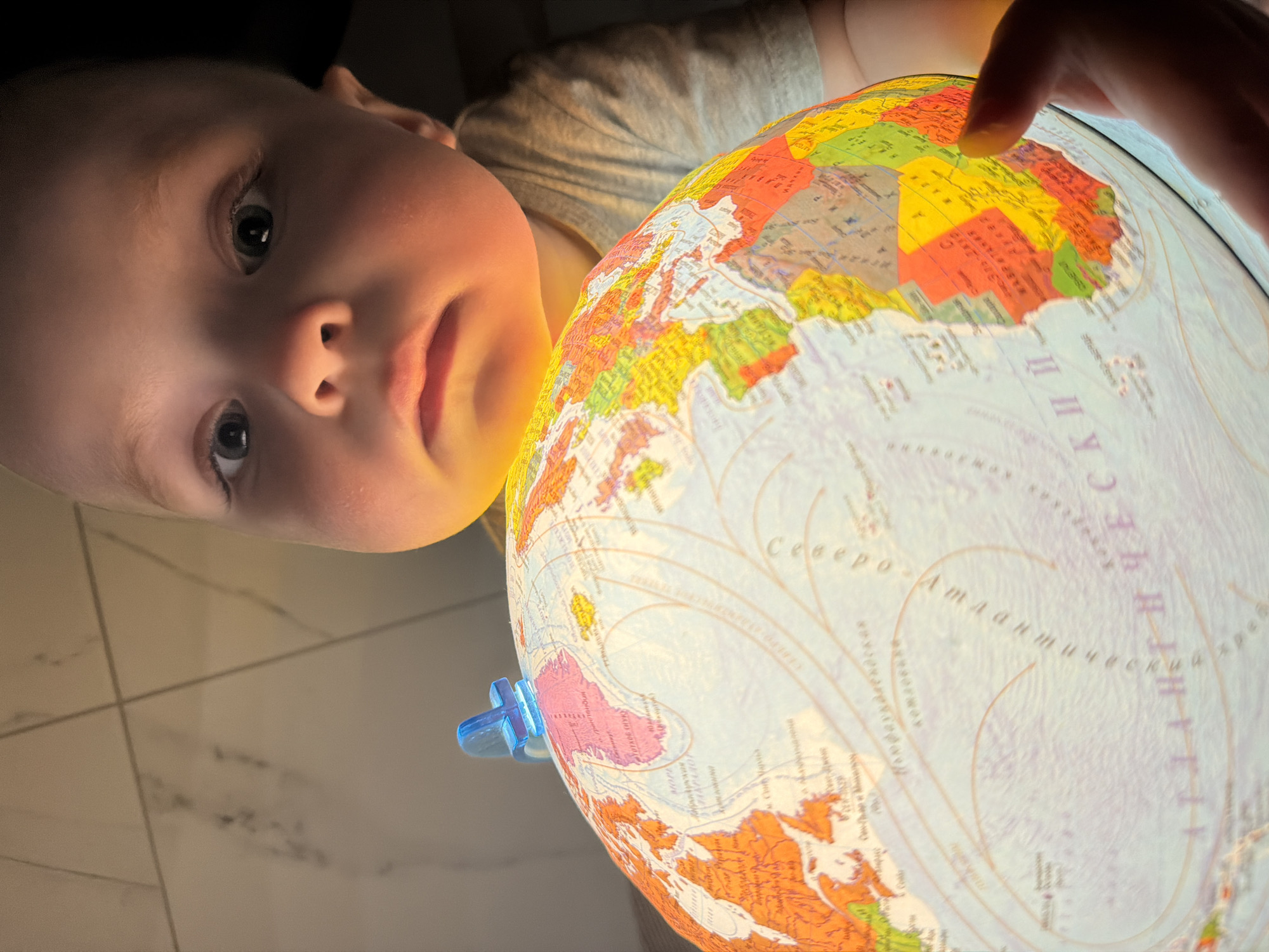 A close-up, low-angle shot shows a young child looking at a lit globe. The child is pale with light eyes and is wearing a light-colored shirt. The globe, brightly lit, displays a map of the world with distinct colors delineating countries and continents. Text in Cyrillic is visible on the globe. A finger touches the globe, possibly indicating a location. The background is a tiled floor and a dark space behind the child. The specific location of Votkinsk, Russia, is not directly identifiable within the visual content of this image.