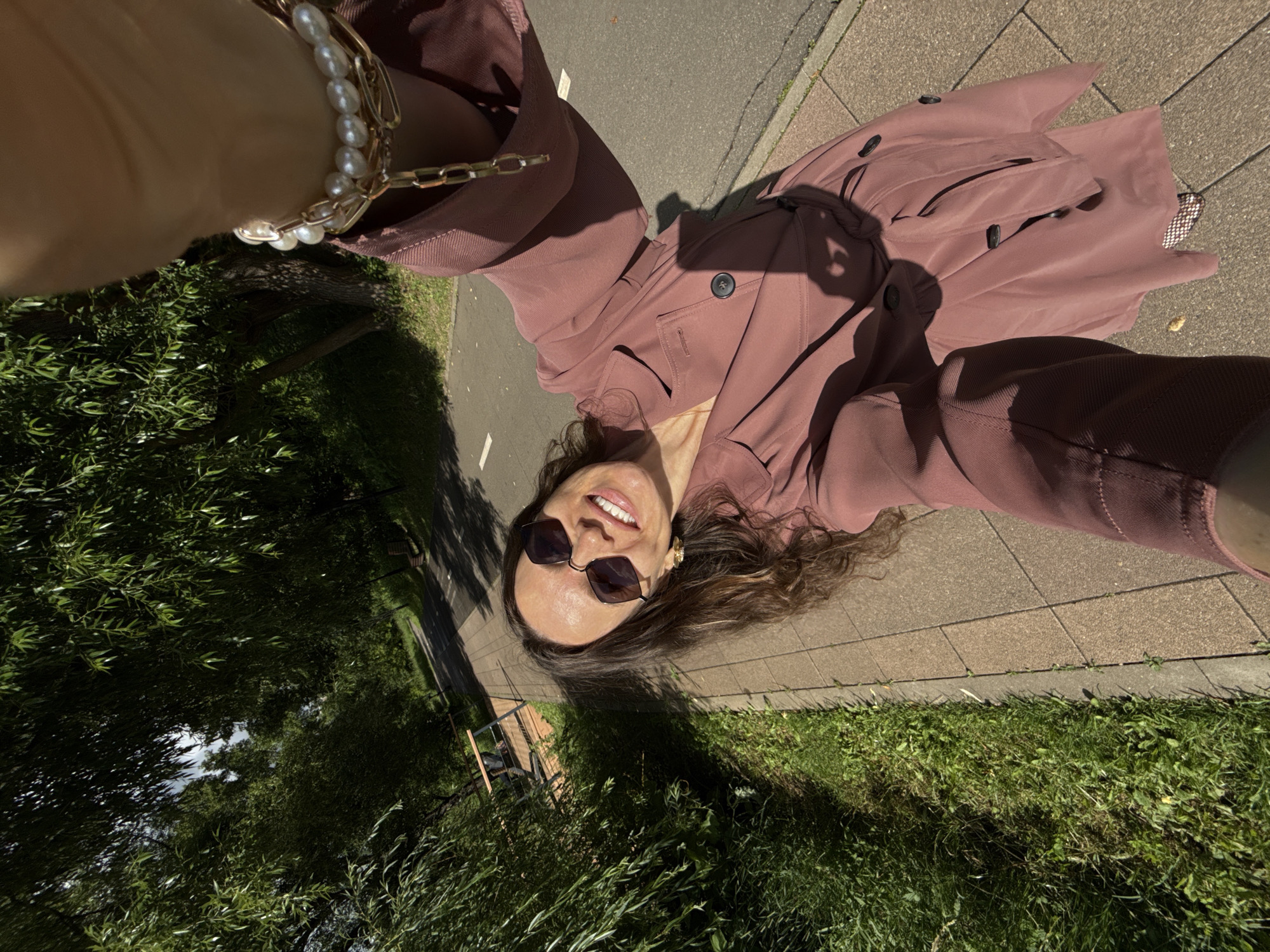 This is a first-person perspective selfie of a woman, likely in Moscow, Russia. She is wearing a mauve trench coat and sunglasses, and a pearl bracelet is visible on her wrist. The woman is smiling broadly, suggesting a happy or cheerful emotion. The setting appears to be outdoors, with a paved walkway and grass visible. Lush green trees with dense foliage are to the left of the frame, and a glimpse of a paved path or road can be seen to the right. The sunlight casts strong shadows, indicating it is daytime, likely a sunny day. The overall impression is one of a casual, pleasant outdoor excursion. There is no visible text in the image.