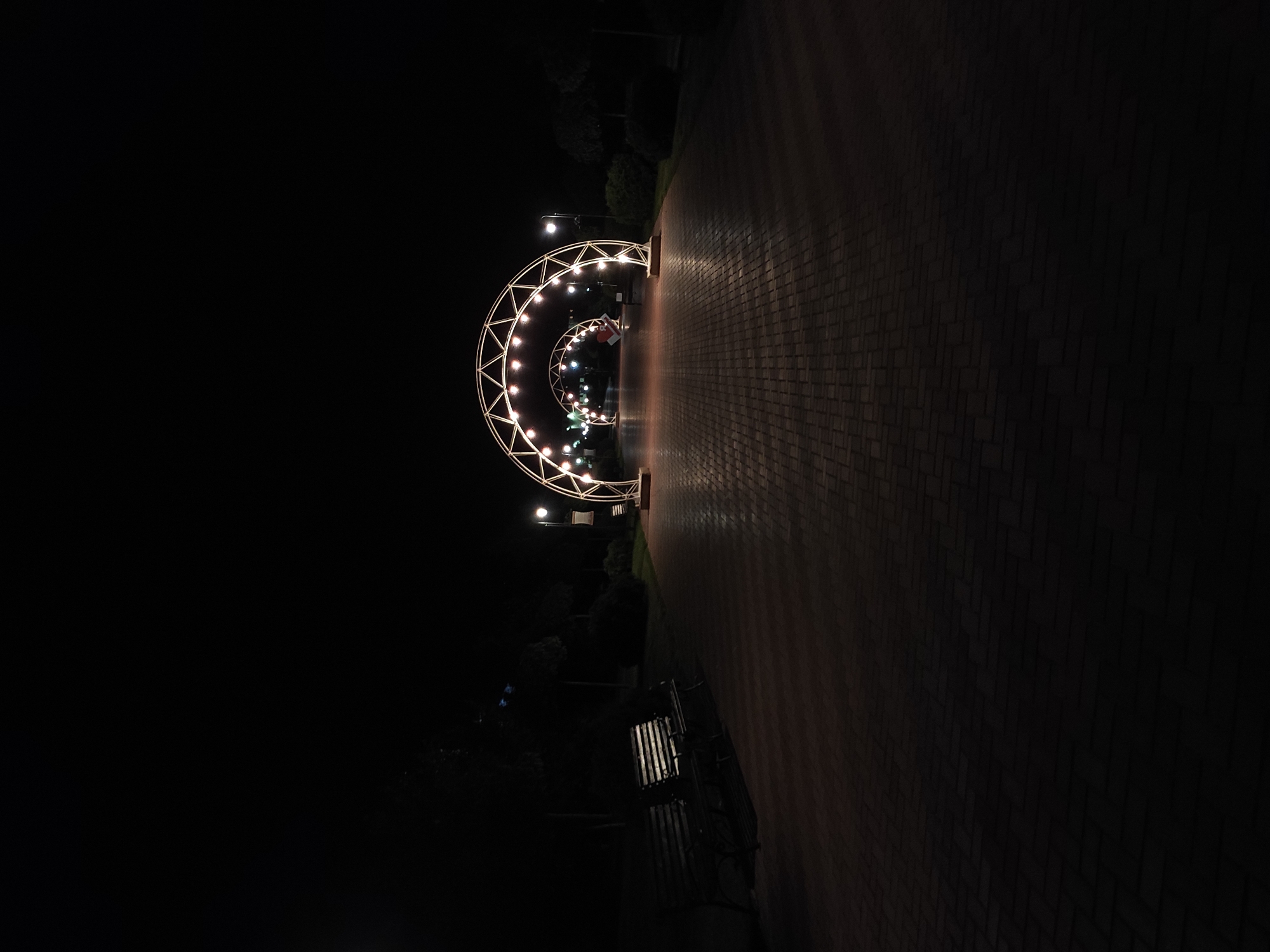 The image shows a nighttime scene in a park. A brick pathway stretches into the distance, lined with illuminated, arched structures adorned with lights. A park bench is visible on the left side of the pathway, and trees and foliage are vaguely discernible in the background. The overall lighting is dim, with the illuminated arches providing the primary source of light. The location is Unknown, Unknown.