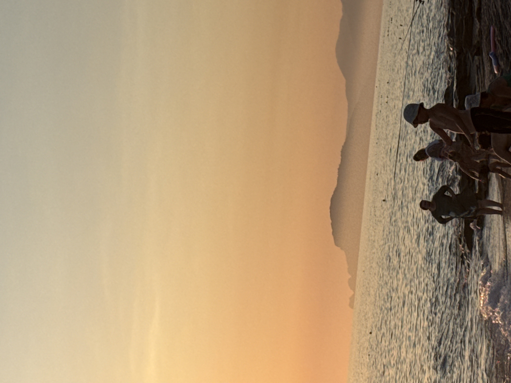 The image depicts a sunset scene at a beach in Alanya, Turkey. The sky is a gradient of pale yellow to soft orange, with a hazy orange glow illuminating the horizon and casting a warm light on the scene. In the distance, silhouetted mountains form a jagged line against the sky. The sea is calm with gentle waves lapping at the shore, reflecting the warm light. A rocky shoreline is visible in the foreground, with some people gathered near the water's edge.
In the immediate foreground, several individuals are present. On the right, a young boy wearing a light blue bucket hat stands facing left, looking out towards the water. He is bare-chested and wearing dark shorts. Next to him, another child, also wearing a light blue bucket hat, is partially visible. To the left of the boy, a group of people are interacting near the water. One person, wearing a dark, possibly green, dress, stands with hands on hips, facing the viewer. Another individual, partially obscured, appears to be a woman in a light-colored garment. Further left, another person can be seen in the water. Several fishing rods are set up along the shoreline, with lines extending into the sea, suggesting recreational fishing. The overall atmosphere is serene and captures a moment of leisure by the Mediterranean Sea.