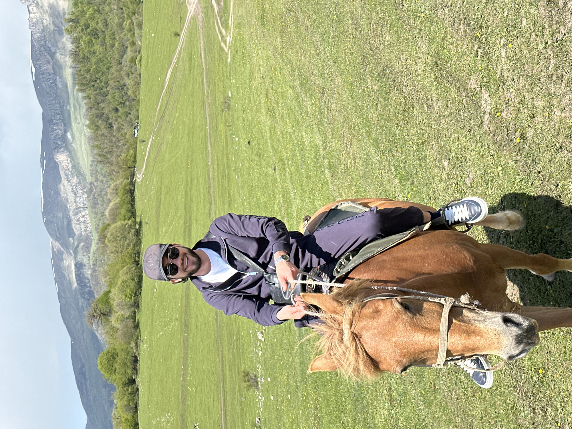 A man rides a horse in a grassy field with mountains in the background, likely in the area of Teghut, Armenia. The man wears a grey baseball cap, sunglasses, a white t-shirt, and a navy blue zip-up jacket and pants. He also wears white and dark grey sneakers. The horse is light brown with a blonde mane and tail. The field is green with some patches of yellow flowers, and there are dirt tracks visible in the distance. The mountains in the background have some patches of snow on their peaks.