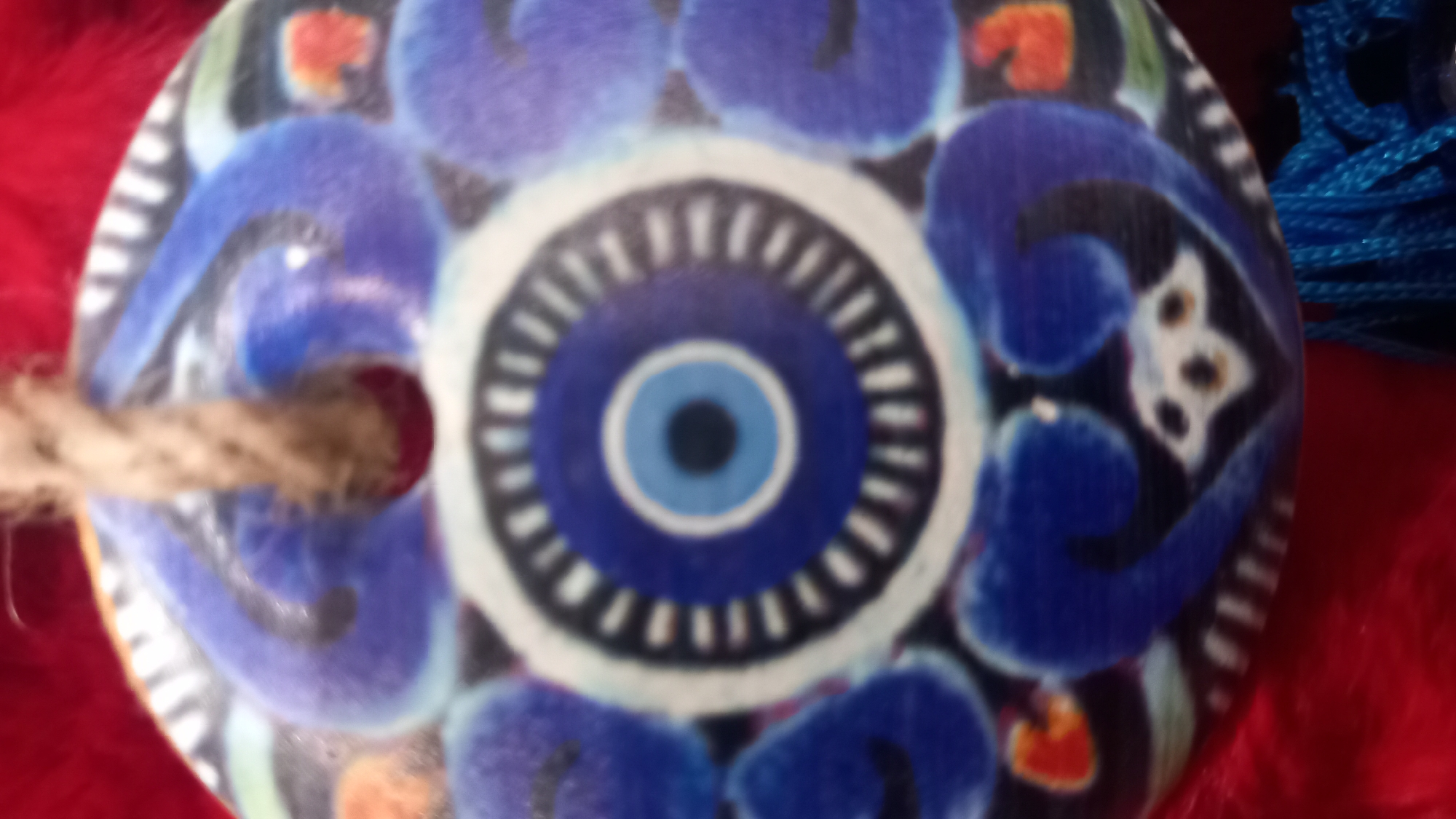 This is a close-up, vertical, slightly blurry, photograph of a circular, decorative object hanging against a red, furry background. The object is made of ceramic and features a traditional "evil eye" design. The design consists of concentric circles in various shades of blue, white, black, and teal. The center is a dark blue pupil within a lighter blue iris, surrounded by rings of black, white, and teal. Elaborate, swirling patterns in dark blue and lighter blue adorn the outer edges of the ceramic piece, interspersed with small orange and green accents. A natural fiber rope loop passes through a hole at the top of the object, suggesting it is an ornament or pendant. A blue tassel hangs from the bottom of the object. The object appears to be a traditional Greek evil eye charm, commonly found in tourist shops and homes throughout Greece, including the island of Kos. Given its appearance and common use, this object is likely a decorative piece or a talisman meant to ward off bad luck.