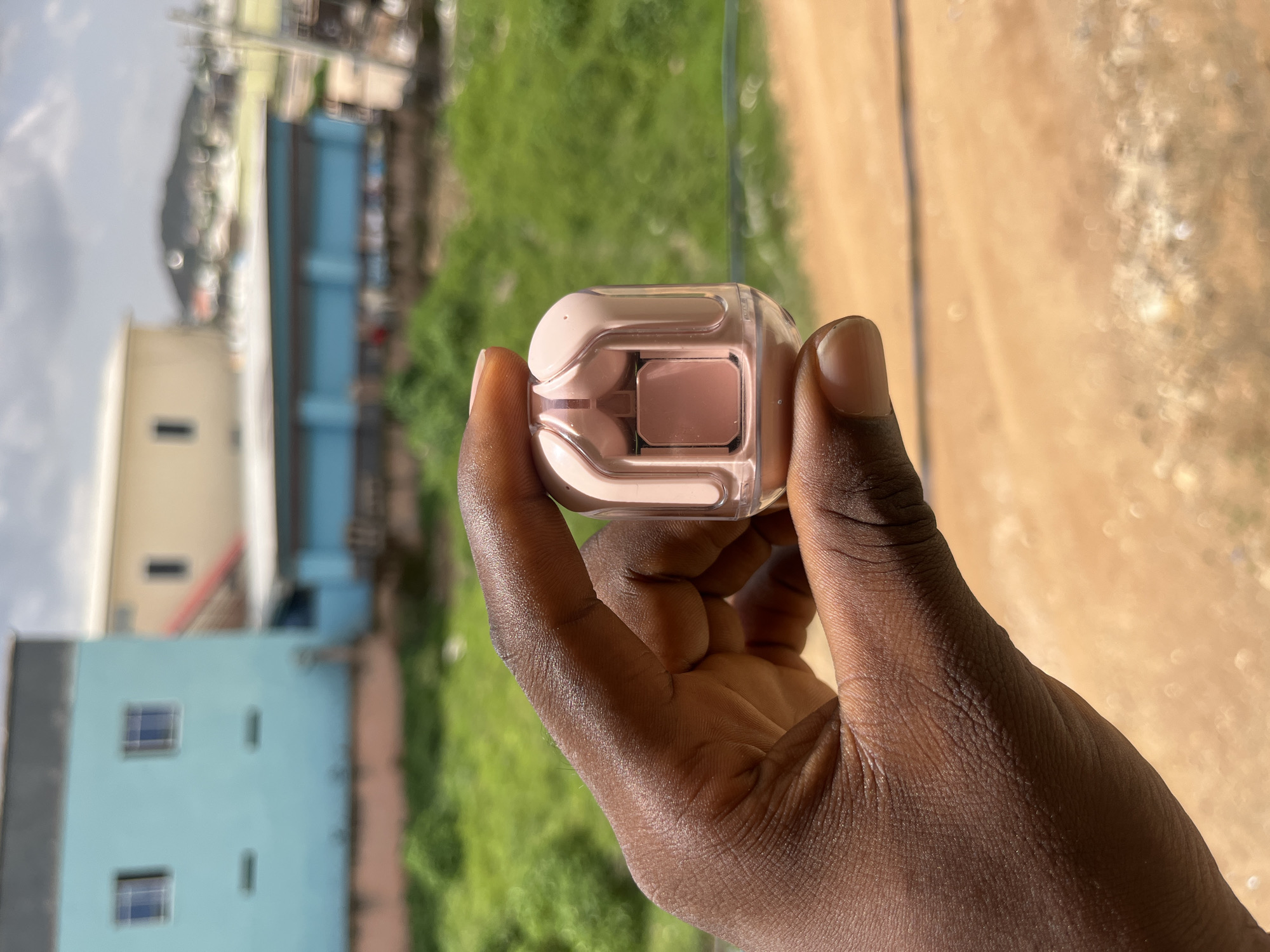 The image shows a close-up of a person's hand holding a light pink and clear wireless earbud case. The hand is dark-skinned, and the person is holding the case in a way that suggests they are examining it or showing it to the camera. The background is blurred but appears to be an outdoor setting with buildings, green foliage, and a dirt path or road. The lighting suggests it is daytime, likely sunny. The overall impression is casual and everyday. There is no visible text in the image. The setting is Gura Top, Nigeria.
