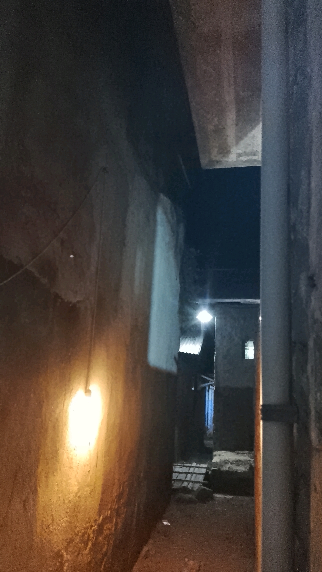 The image depicts a narrow, dimly lit alleyway at night in Ujungrusi, Indonesia. A light bulb hangs from a wire on the left wall, casting a warm glow on the rough, textured surface. The alley is flanked by buildings on both sides, one with a corrugated metal roof visible. In the distance, a street light illuminates the path, revealing more structures and some scattered debris. A large pipe runs vertically along the right side of the alley. The overall impression is one of a somewhat impoverished or older urban environment.