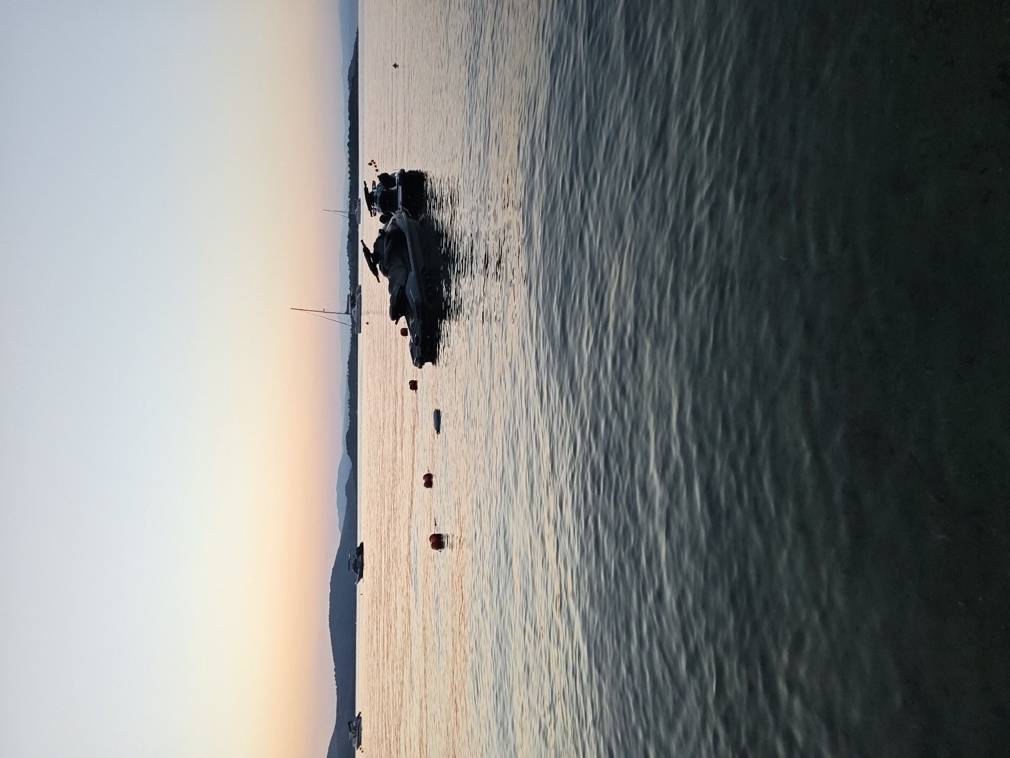 A serene seascape at sunset is captured from the water's edge, likely within Sithonia, Greece, given the characteristic rolling hills and tranquil waters often found in the region. The sky transitions from a pale, clear blue at the zenith to soft hues of peach and orange near the horizon, indicating the sun is low. In the mid-ground, a calm body of water reflects the sky's warm tones, with gentle ripples disturbing the surface.
Several objects are dotted across the water. Two jet skis are moored together in the right-center of the frame, their dark silhouettes stark against the glowing water. To their left and further into the distance, a larger boat with a cabin is visible, also at anchor. Further to the left, a smaller boat is seen. A line of orange buoys marks a boundary or pathway in the water, stretching from the left side of the frame towards the jet skis. In the far distance, a long, dark landmass with undulating hills stretches across the horizon, fringed by a faint purple haze. The water in the foreground is darker and more opaque, with subtle patterns created by the waves, suggesting a shallower area near the viewer. No people are visible in the frame.