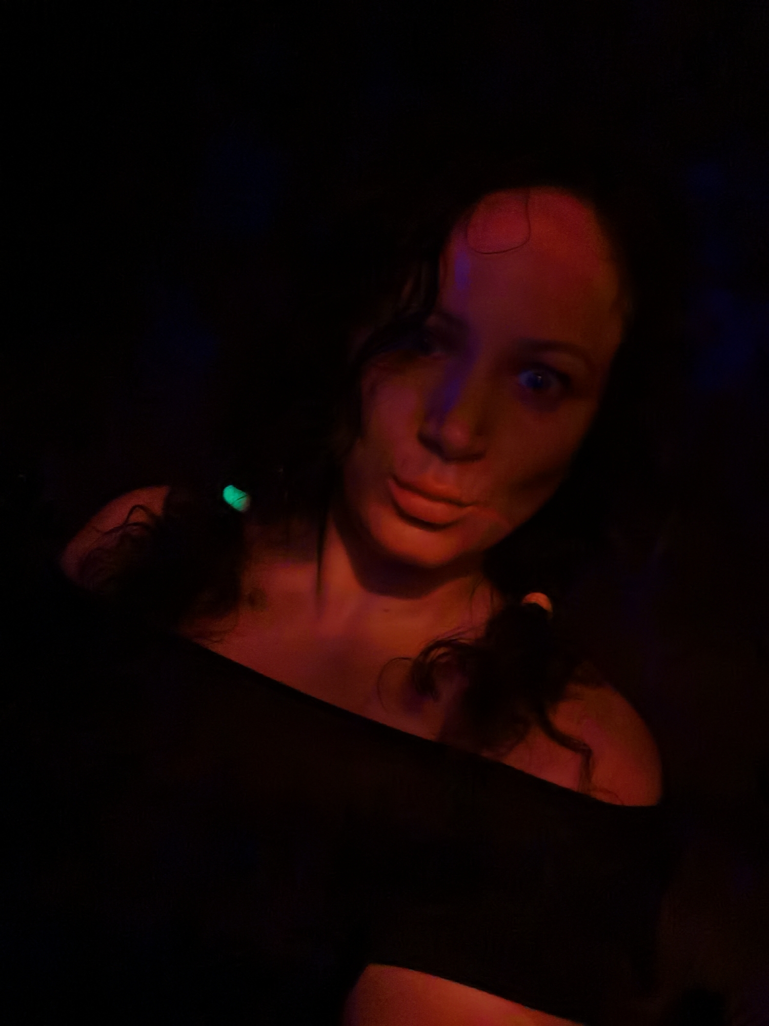 A woman is seen in a dimly lit, red-hued environment. Her face, illuminated by the faint light, shows a curious expression. She has dark, curly hair, with a green light attached to one side and an orange light on the other. She is wearing a black off-the-shoulder top. The location appears to be in the city of Unknown, Unknown, but specific details are not discernible due to the low lighting conditions.