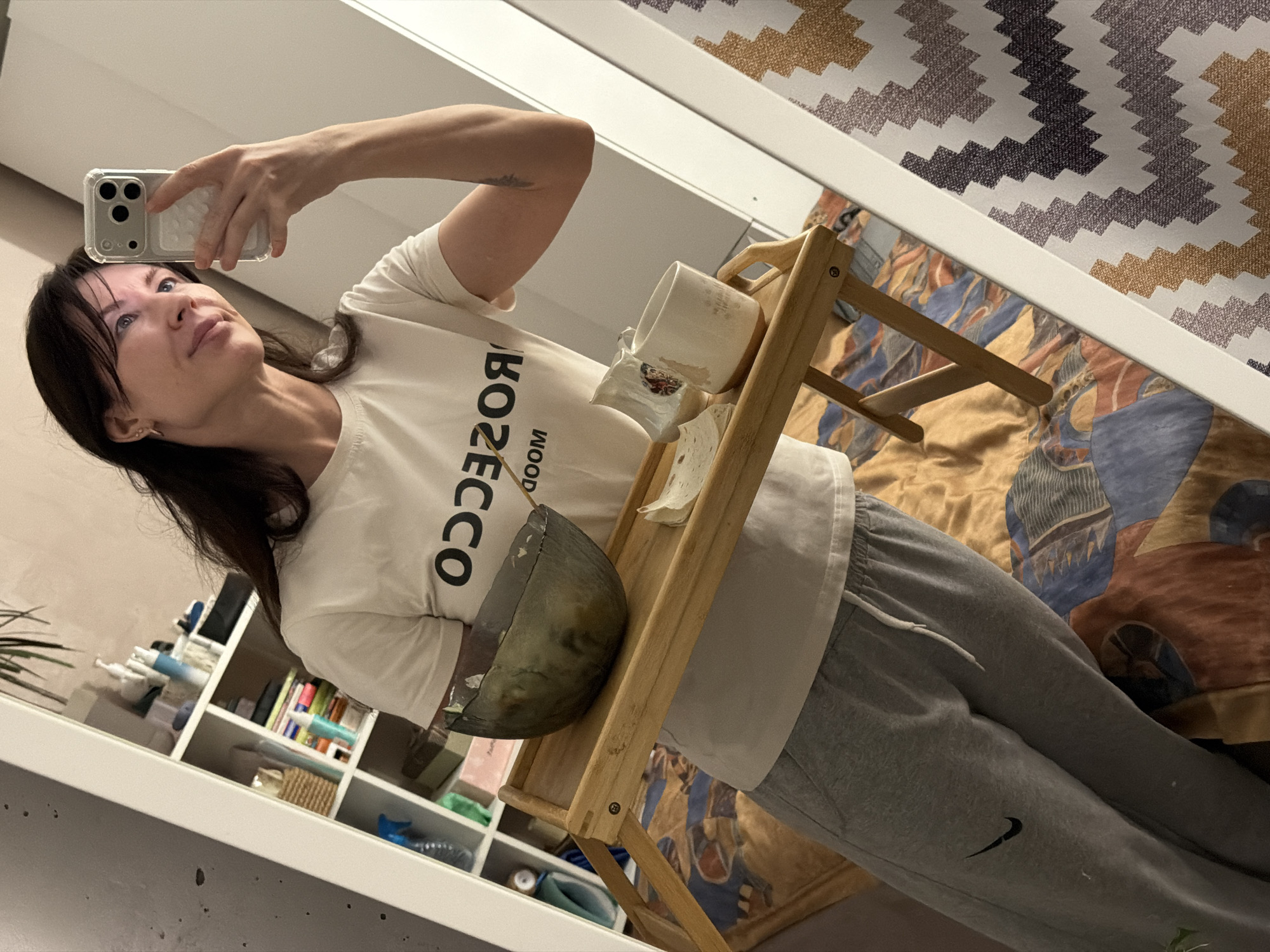 In an interior setting in Perm, Russia, a female individual is observed taking a mirror selfie. She has dark hair and is wearing a white t-shirt with "PROSECCO DOOM" printed in reverse, and grey Nike sweatpants. Her right hand holds a light-colored smartphone with a clear case, positioned to capture her reflection. Her left arm supports a light-colored wooden breakfast tray. On the tray, from left to right, are a dark-tinted glass bowl containing a light-colored food item, a small white dish holding what appears to be flatbread, and a white ceramic mug with visible stains. The background, reflected in the mirror, reveals a white modular shelving unit filled with various personal items, including bottles, baskets, and a plant. Below the tray, a bed is visible with a patterned comforter featuring geometric shapes in warm and cool tones. To the right of the subject, a wall or door with a white trim and a textile or wallpaper with a zigzag pattern in brown, purple, and white is reflected.