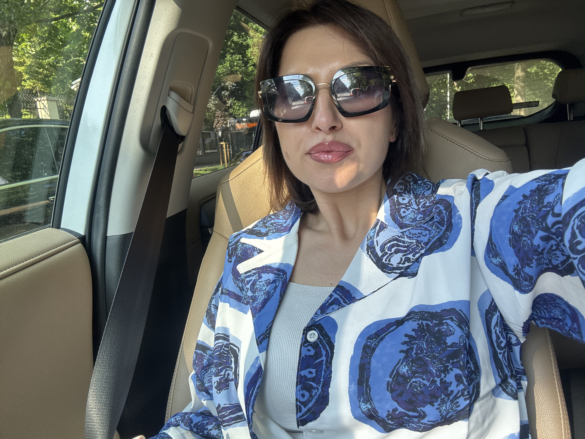 A middle-aged woman with short brown hair and large sunglasses is seated in the passenger seat of a car, taking a selfie. She is wearing a white shirt with a blue floral pattern and a light-colored tank top underneath. The interior of the car is visible, with beige leather seats and a black seatbelt. Outside the car window, trees and other vehicles can be seen, suggesting an urban setting. The lighting indicates it is daytime, possibly late afternoon due to the long shadows. The image appears to have been taken while the car was parked or moving slowly, given the clear focus on the woman. The background details, such as the fence and a building with a sign that appears to read "TOYOTA", suggest the location is in Sofia, Bulgaria, specifically on a street lined with trees and parked cars.