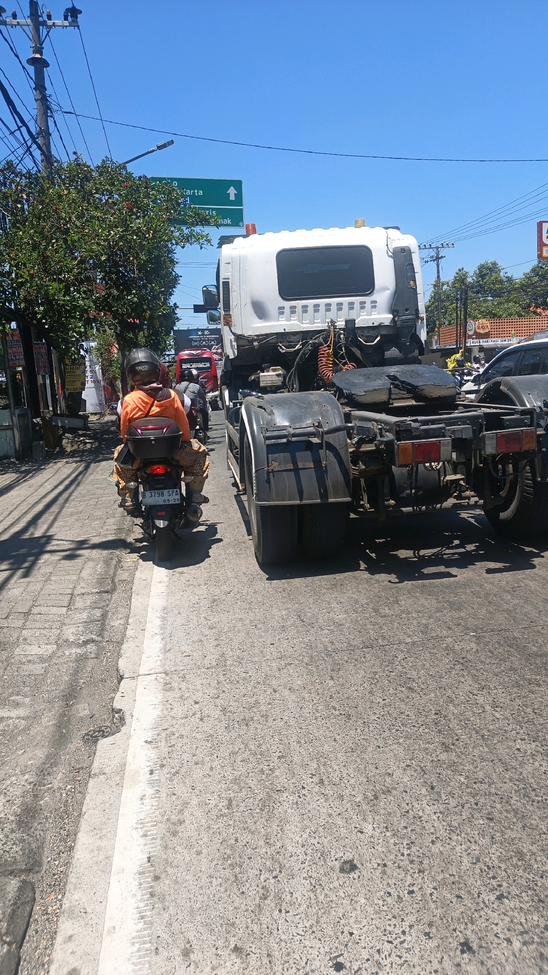 An exterior, eye-level shot captures a sunny daytime scene on a paved road in Genuk Barat, Indonesia. The foreground is dominated by the rear of a white semi-trailer truck, with its large black tires and chassis prominently displayed. To the left, a person wearing an orange jacket and patterned pants rides a motorcycle, also viewed from behind. The motorcycle has a black top box and a license plate that reads "B 3798 SPA 09-29". The road is bordered by a concrete sidewalk and a patch of gravel. In the background, a green directional sign pointing upwards reads "Jakarta" and "Banyuwangi". Trees line the left side of the road, and power lines crisscross the bright blue sky. Other vehicles, including another truck and cars, are visible further down the road, suggesting traffic. The overall impression is of a typical urban street scene during daylight hours.