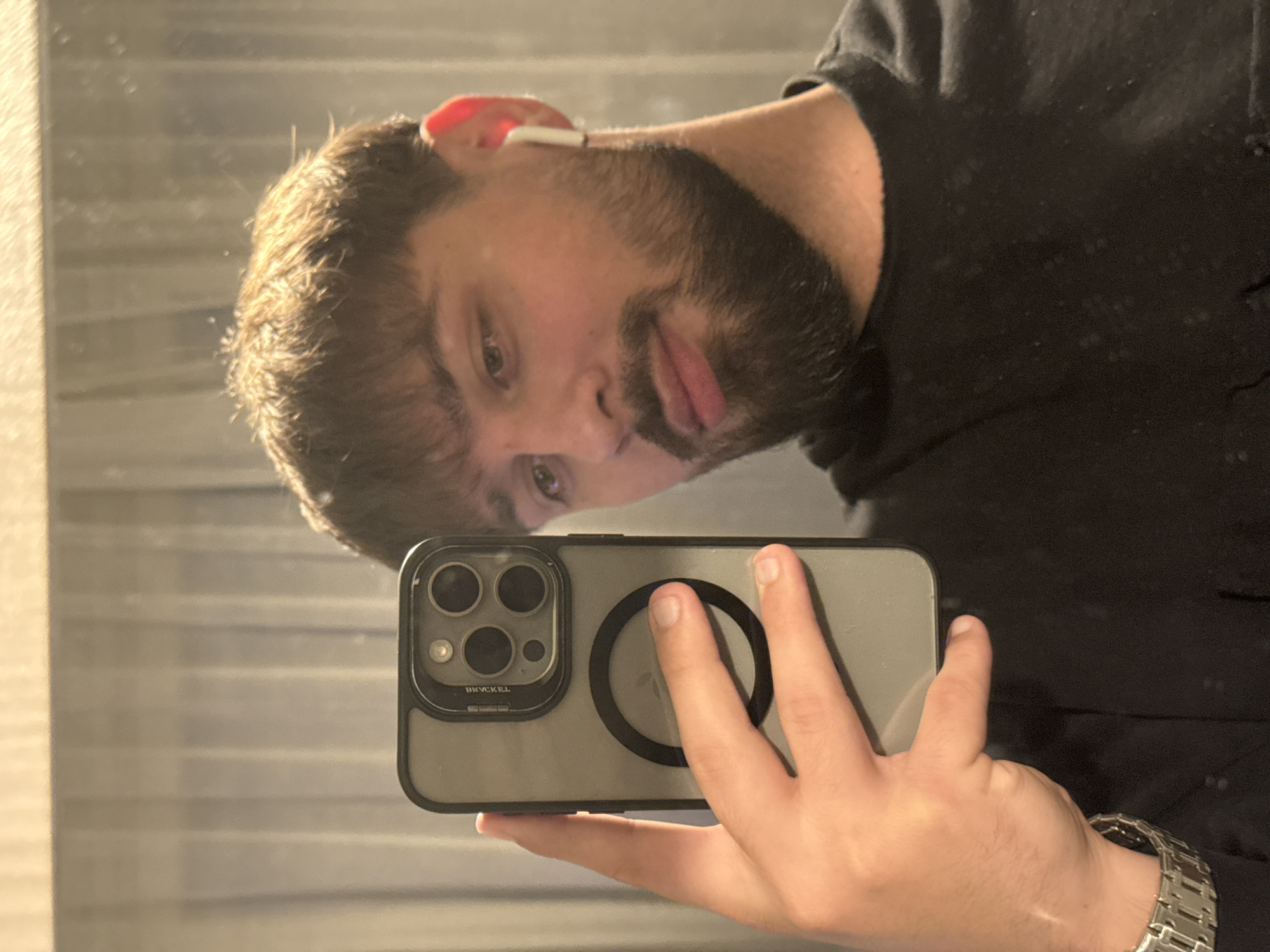 A man with a beard and short hair is taking a selfie in a mirror with his iPhone, which has a black case with a circular magnetic attachment on the back. He is wearing a black shirt and a metal watch. He has an earbud in his right ear. The location is not evident in this image. It is not possible to determine if the location is in Varna, Bulgaria or anywhere else.