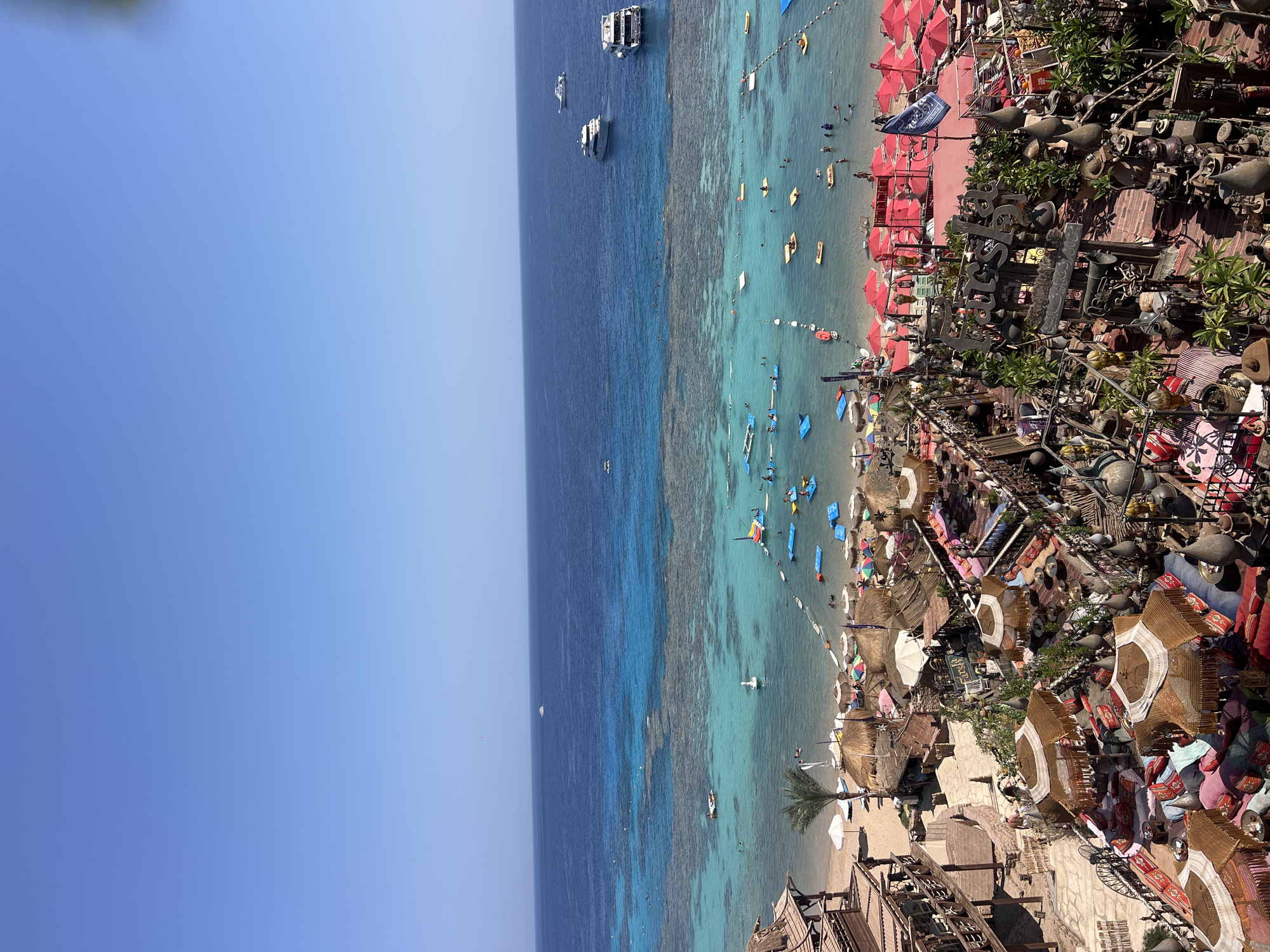 This is an aerial, wide shot of a beach resort in Sharm El Sheikh, Egypt. The scene depicts a vibrant, tiered outdoor seating area in the foreground, featuring numerous low tables, cushions, and ornate umbrellas. The seating area overlooks a sandy beach with thatched-roof cabanas and rows of red umbrellas. Beyond the beach, the turquoise water transitions to a deep blue as it meets the horizon. Coral reefs are visible beneath the clear water, and various boats, including yachts and smaller vessels, are anchored or cruising in the sea. Several people are visible on the beach, in the water, and on the various floating platforms and watercraft. The sky is a clear, bright blue. The resort appears to be a popular tourist destination, likely a beach club or restaurant, with a lively atmosphere.