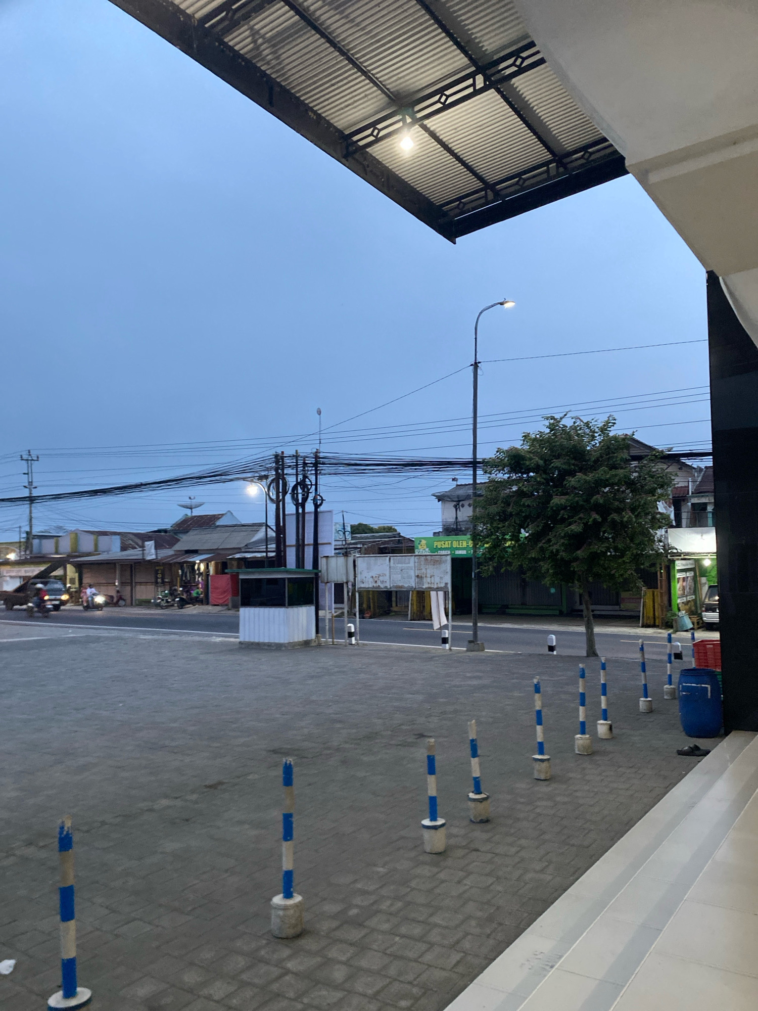 The image depicts an outdoor scene in Karangluhur, Indonesia, likely during twilight or early morning given the soft, even lighting and the overcast sky. In the foreground, a paved area with a textured, grey stone surface is marked by a series of striped bollards – white with blue bands, standing on concrete bases. To the right, a portion of a modern building's facade, painted white, is visible. Beyond the paved area, a street runs horizontally, with a row of low-rise buildings lining its opposite side. These buildings appear to be shops or small businesses, with some displaying signs and wares. A prominent tree with dense green foliage stands near the street, partially obscuring the buildings behind it. Overhead, a network of electrical wires crisscrosses the sky, supported by poles. A street lamp with a lit bulb is positioned over the street. In the middle ground, a small, enclosed booth or kiosk with white paneling and dark-tinted windows is situated near the street. To the far left, a dark-colored vehicle and a motorcycle are visible on the street, suggesting some traffic. The overall atmosphere is quiet and subdued, indicative of a less busy time of day.