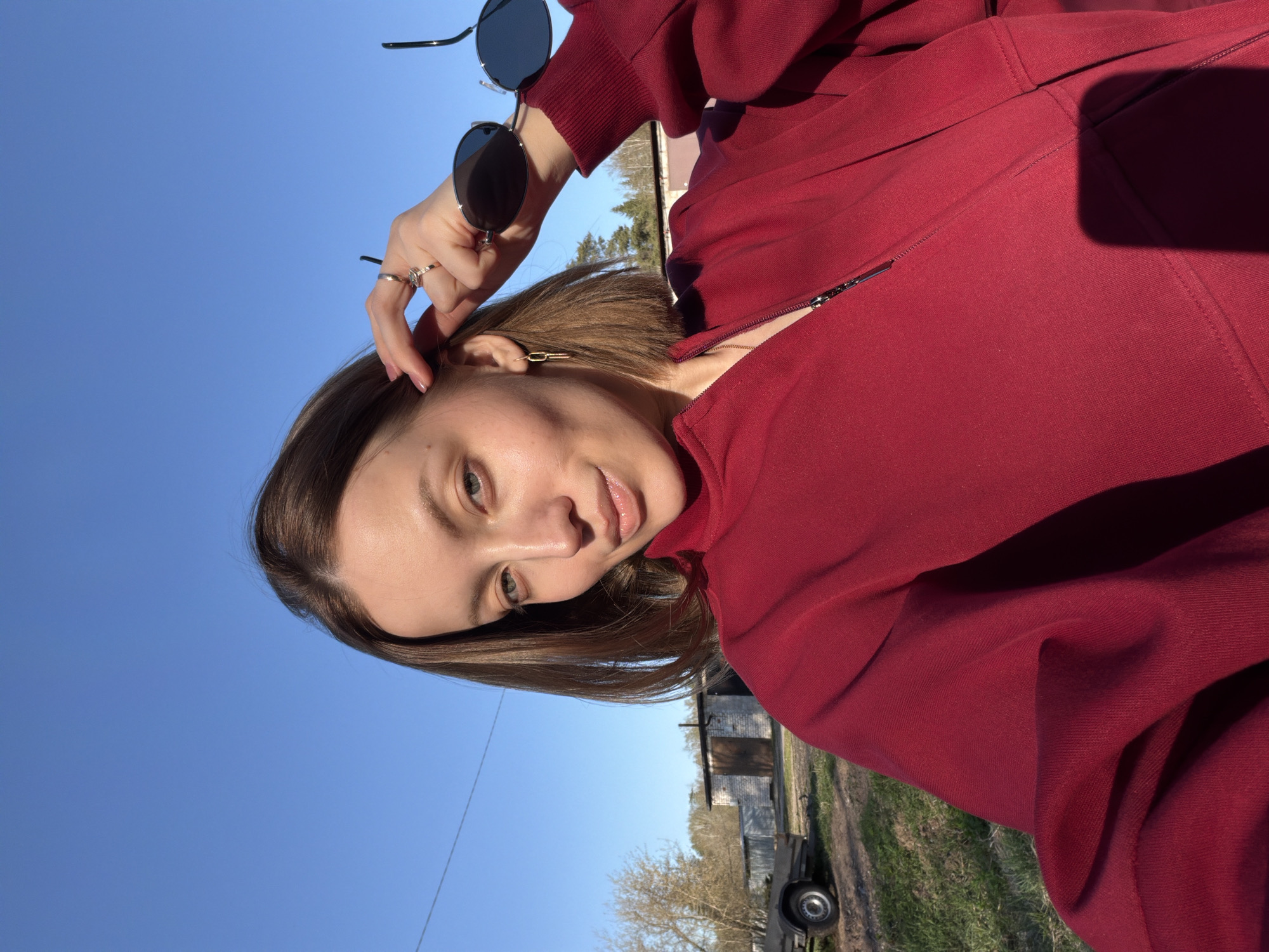 A young woman in a red jacket is standing outside in Reshetnikovo, Russia. She is holding sunglasses in her left hand and touching her hair with her right. In the background there is a blue sky, a grassy area, and a structure.
