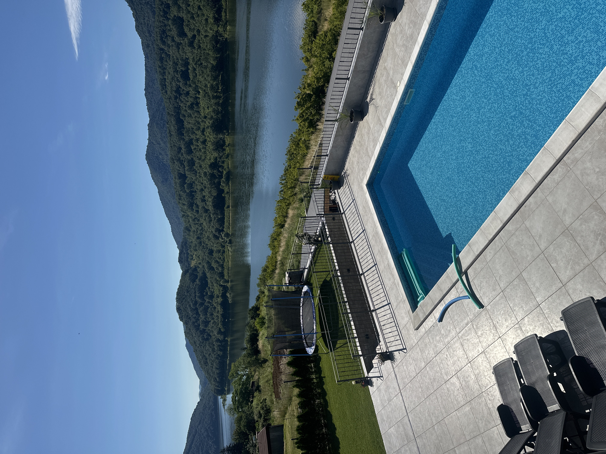 This is a high-angle, wide shot of a swimming pool area with a natural body of water and forested hills in the background under a clear blue sky. In the foreground, a bright blue, rectangular swimming pool with tiled edges is filled with water. Several dark-colored lounge chairs are arranged on the grey tiled patio surrounding the pool. Two green pool noodles are floating in the pool, and a blue hose lies across the tiles. A black metal fence separates the pool area from a grassy lawn with a trampoline in the middle. Beyond the lawn, a dark body of water, likely a lake or river, stretches into the distance, bordered by lush green, tree-covered hills. A small building and some fencing are visible on the left side of the frame. The scene appears to be a residential property.
