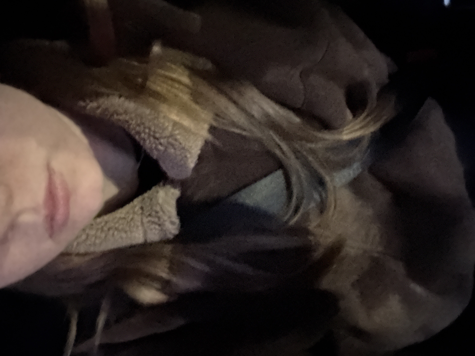 A close-up, low-light image captures a person, likely female, resting or sleeping. The subject's face is partially visible on the left side of the frame, showing a light-skinned cheek, part of the nose, and lips. Long, blonde hair flows across the upper and central parts of the image. The person is wearing a dark-colored jacket or coat with a prominent, light-colored, fluffy, possibly sherpa, lining around the collar. The head is tilted, suggesting a relaxed posture. The background is mostly dark and indistinct, indicating a nighttime setting or a dimly lit indoor environment, possibly within a vehicle given the intimate and confined feel. No specific activity beyond resting is discernible. The overall mood is peaceful. There is no visible text in the image. The location context is Chaikovsky, Russia.