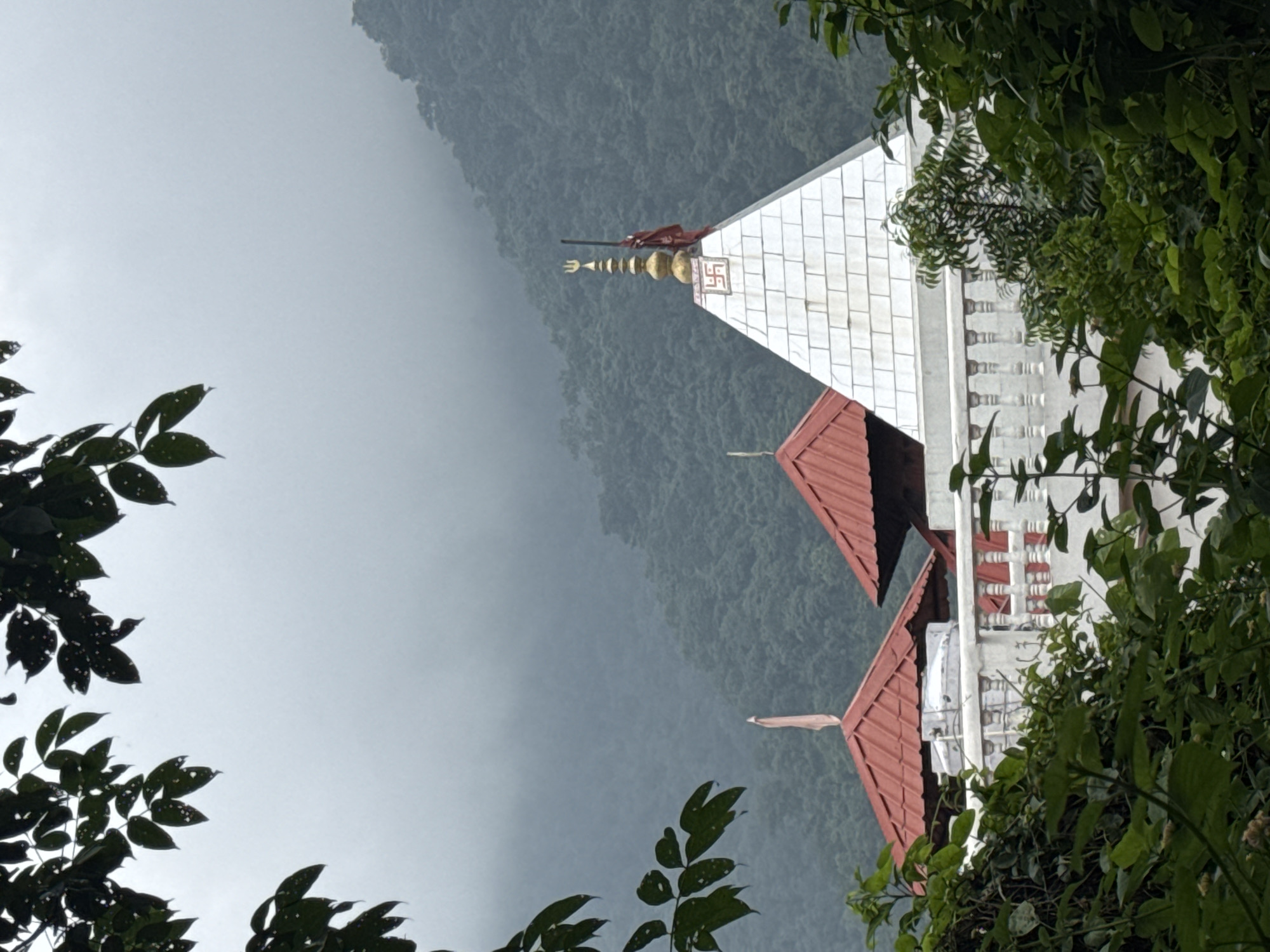 The image depicts a Hindu temple structure in Rishikesh, India, nestled amidst lush green hills shrouded in mist. The temple features a white, pyramid-shaped spire adorned with a golden pinnacle, a trident symbol, and a Swastika emblem. Adjacent to it, a smaller structure with a red tiled roof is visible, along with a white balustrade. Green foliage in the foreground partially obscures the view of the temple. The overall atmosphere is serene, with the misty mountains suggesting a cloudy or rainy day.