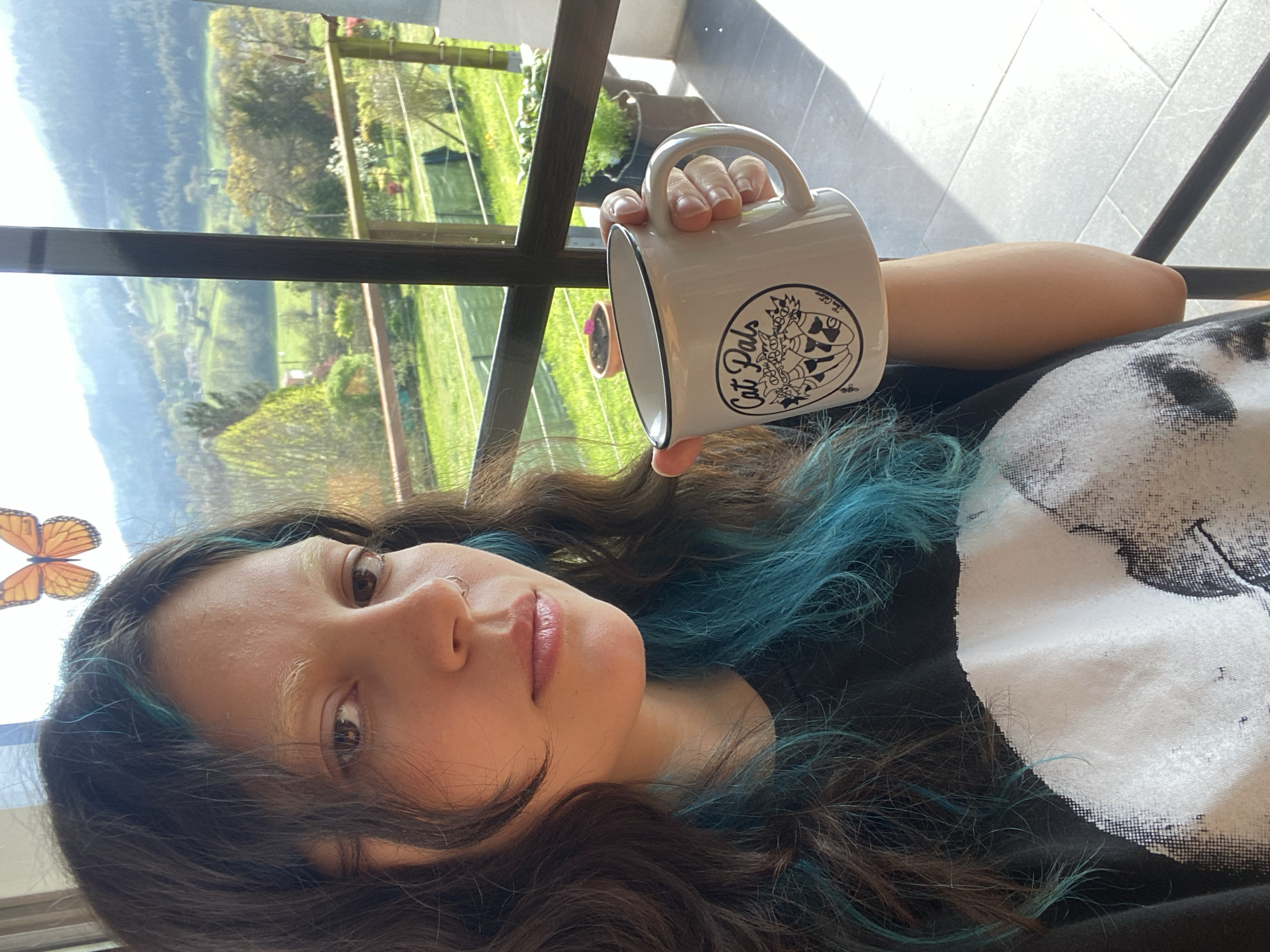 The image is a selfie taken indoors, likely on a balcony or near a large window, with a clear view of a landscape typical of Villaviciosa, Spain, characterized by green rolling hills and distant mountains under a sunny sky. A woman with long, dark brown hair with blue highlights is holding a white mug with the words "Cat Pals" printed on it. She is wearing a dark T-shirt with a large white print. Her skin tone is fair and she has a nose ring. The photo is taken from a low angle. A decorative butterfly is on the left side of the image. The environment outside suggests a rural or suburban area near Villaviciosa, based on the landscape.