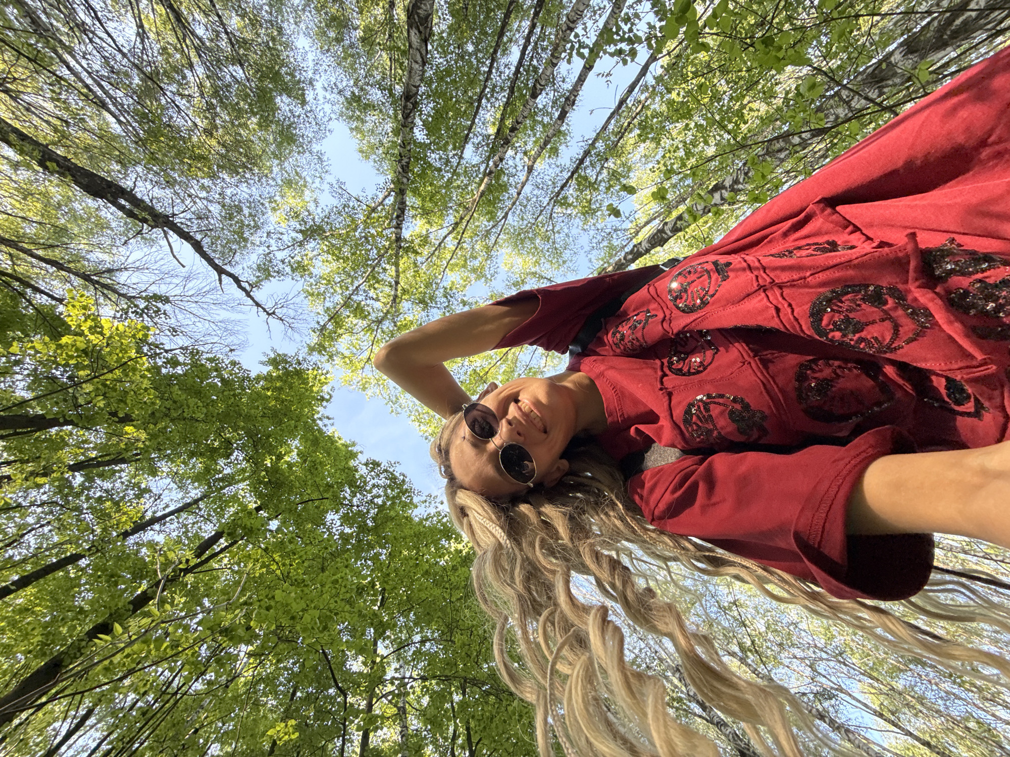 A low-angle, upward-facing view captures a woman posing against a backdrop of a forest canopy. The woman wears round sunglasses and a red top with embellished details. Her long, blonde hair hangs downwards. Above her, green leaves of trees fill most of the frame, with glimpses of blue sky visible between the branches. The location is Шебунино, Russia.