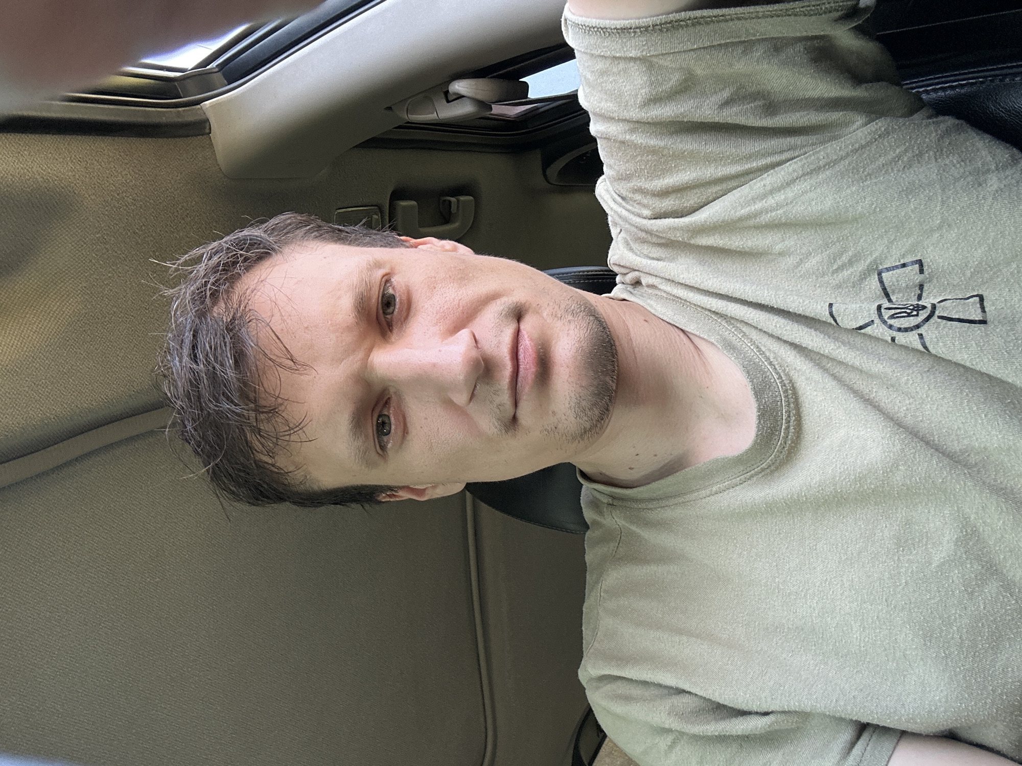 A man is sitting in the driver's seat of a car, looking directly at the camera. He has light brown hair, light skin, and is wearing a light olive-colored t-shirt. The interior of the car is visible, including the ceiling and a portion of the car door. The image is taken from a head-on perspective, with the man's face and upper body filling most of the frame. There is a small, black symbol on the man's t-shirt, to the left of his chest. The lighting appears to be natural daylight, suggesting the photo was taken outdoors. The overall scene is casual and unposed.