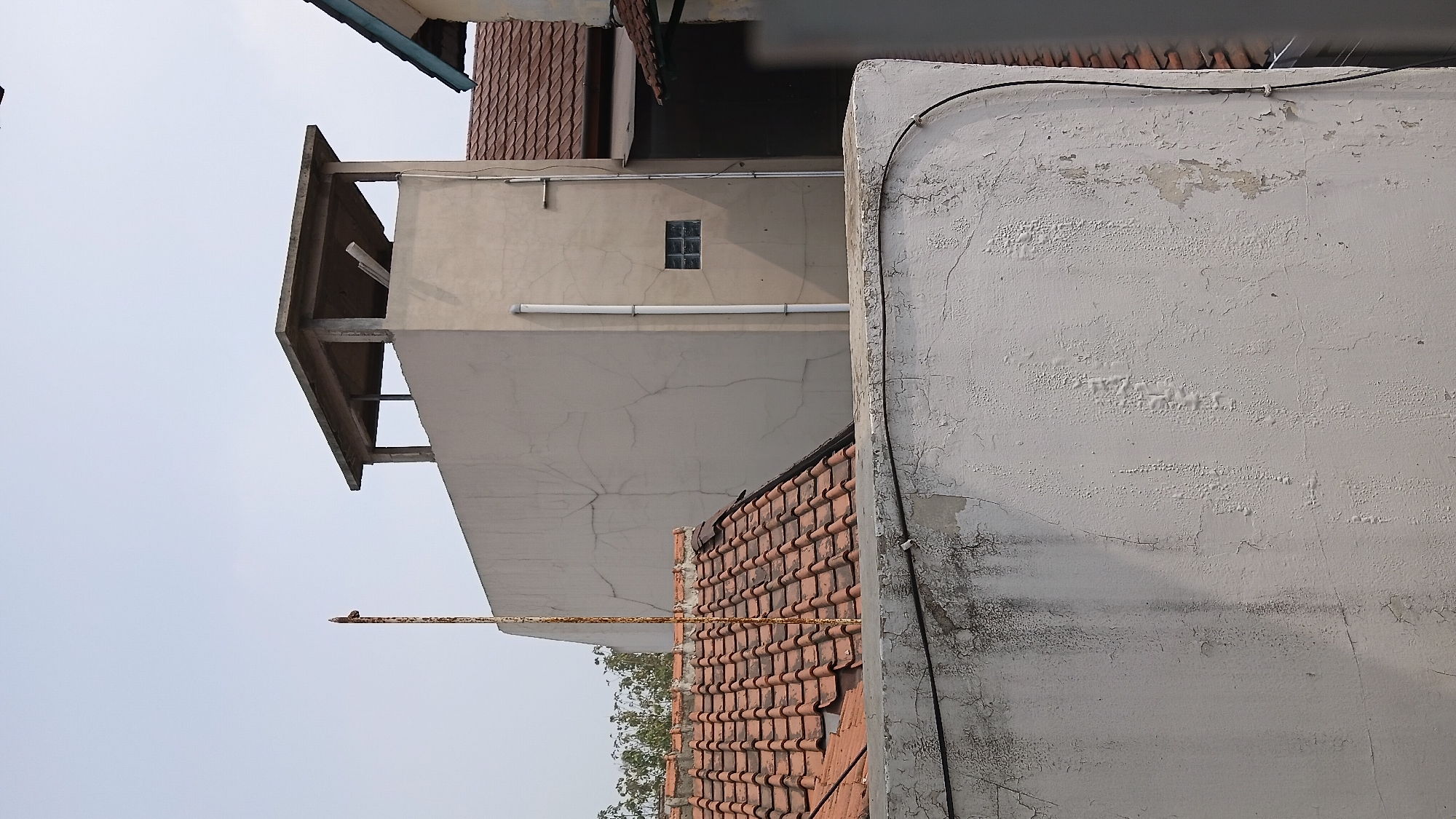 The image depicts a rooftop view in an urban setting, likely a residential area, under a bright sky. In the foreground, a weathered, off-white wall with peeling paint and visible cracks dominates the lower portion of the frame. A black electrical wire is attached to the wall with white clips.
Behind this wall, a section of a terracotta-tiled roof is visible, angled and showing signs of age. A rusty metal pole extends vertically from behind this roof, with a small, horizontal metal piece attached to its top, resembling a rudimentary antenna or support.
Further back, a beige, multi-story building with a flat, unfinished concrete roof structure on top stands prominently. This upper structure has open sides supported by concrete pillars. A single, small, dark-colored glass block window is set into the beige wall of this building. A white vertical pipe runs down the exterior of the building. To the right of this building, another roof with terracotta tiles and a darker sloped roof with a green trim are partially visible, suggesting the presence of adjacent structures. The overall scene conveys a sense of an older urban landscape, possibly within the city of Bandung, Indonesia, characterized by visible wear and tear on the structures.