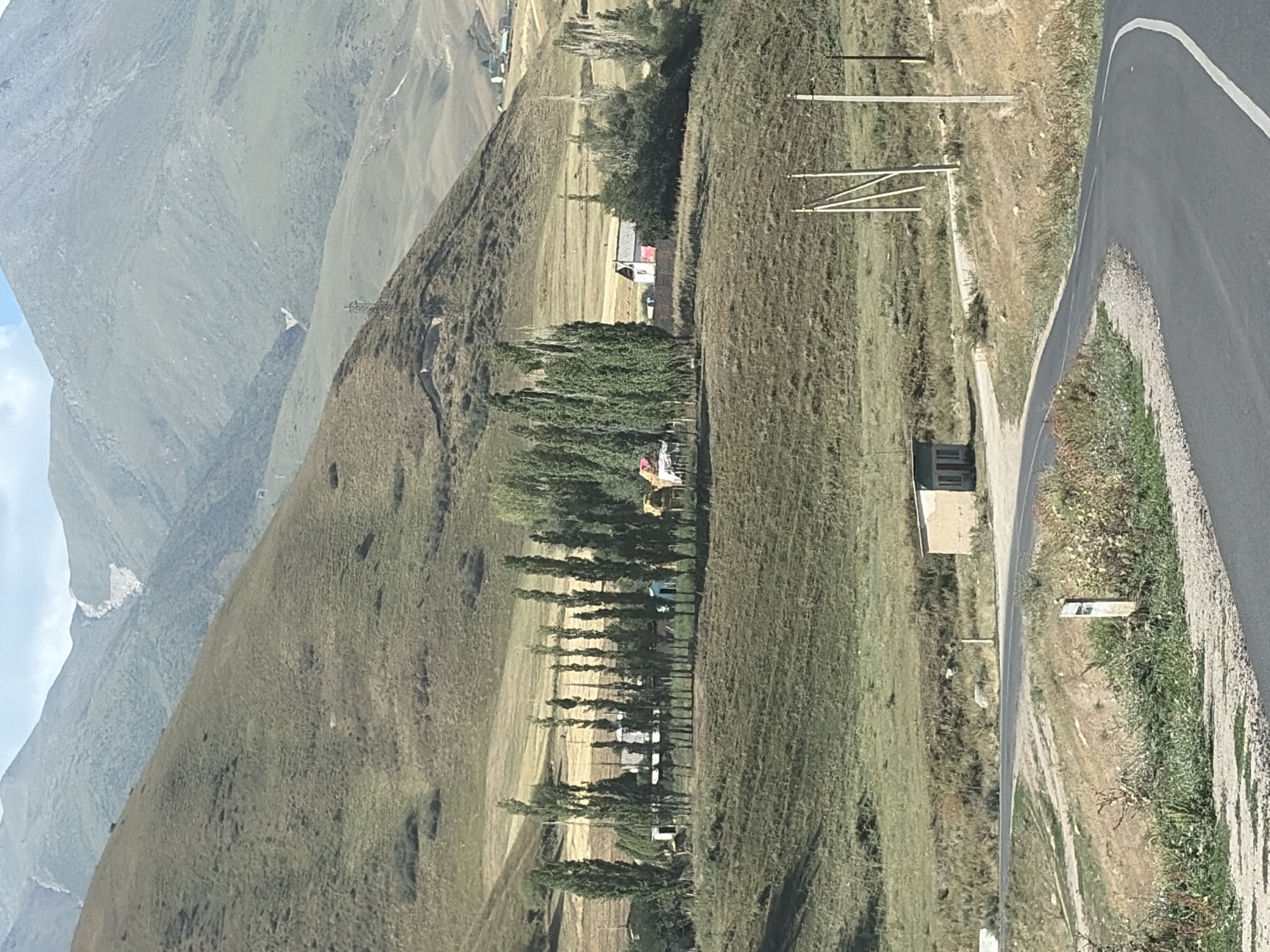 A paved road curves through a rural, mountainous landscape. A small, light-colored building with a dark roof sits to the right of the road in the mid-ground. A line of tall, slender trees stands in a row on a slightly elevated grassy area to the left of the road, with several small buildings and some hay bales visible behind them. The background is dominated by rolling, grassy hills and mountains, with a patch of lighter-colored terrain on one of the larger mountains in the distance. Power poles with wires are visible along the right side of the road in the foreground. The sky is cloudy with patches of blue.