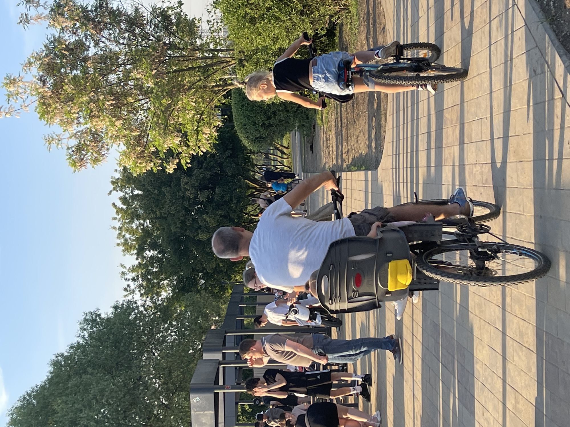 This image captures an outdoor scene in a paved park or recreational area in Moscow, Russia, during what appears to be late afternoon or early evening due to the long shadows.
In the immediate foreground, a bald adult male, viewed from behind, rides a dark-colored bicycle. He wears a white t-shirt and grey shorts. A dark grey child bicycle seat, equipped with a red reflector and a yellow lever, is mounted on the rear of his bicycle. To his right, a young girl, possibly pre-teen, also rides a dark-colored bicycle with blue accents. Her blonde hair is tied in a high ponytail, and she wears a black sleeveless crop top and light blue denim shorts. She is looking back over her left shoulder, towards the area where the adult male is situated. Both cyclists are on a light-colored paved pathway composed of rectangular pavers.
To the left of the cyclists, a group of approximately ten individuals is gathered or walking on the same paved path. Notable figures include a man in a brown polo shirt and blue jeans, standing with hands on his hips and looking towards the right. Nearby, a younger male in a black t-shirt and shorts has both hands covering his face or forehead. Another male in a white t-shirt stands close to a white scooter. In the far left background, multiple dark-colored, open-sided structures with metal frames are visible, consistent with modern park infrastructure such as pergolas or kiosks.
The background is dominated by lush green foliage and numerous trees. On the right, a tall tree with a distinct light-colored, possibly flowering or seed-bearing, canopy stands out. A dense green hedge lines the paved path on the right, beyond which a dirt track is visible. The sky above is a clear, light blue, indicating fair weather. Strong, low-angle sunlight casts long, distinct shadows of the people and trees across the paved surface.