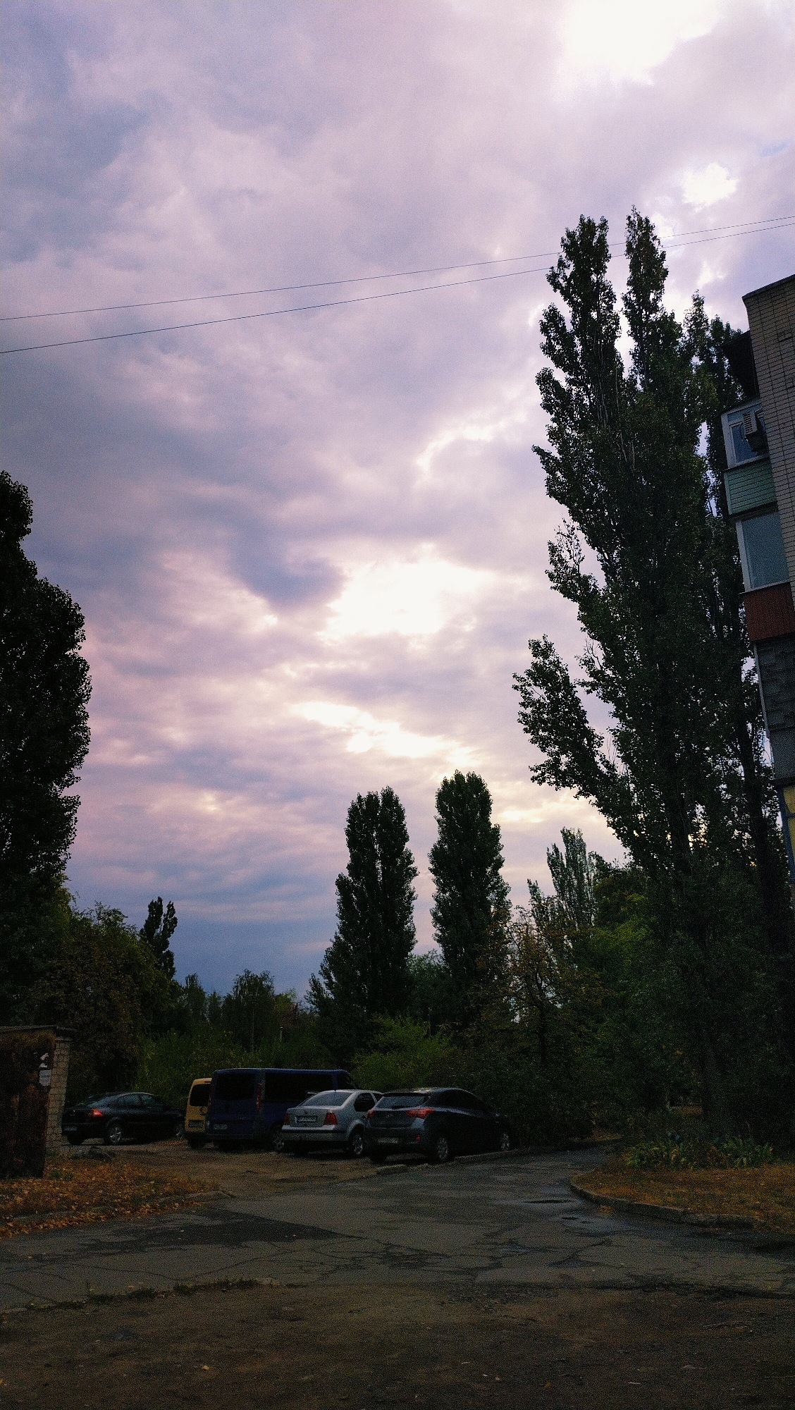 The image captures a street scene in Horishni Plavni, Ukraine, featuring a view of a parking area in front of a multi-story brick building. The sky above is filled with dramatic clouds, tinged with purple and pink hues, suggesting either early morning or late evening. Tall, slender trees with dense foliage line the street and partially obscure the building. Several cars are parked in the lot, including a dark sedan, a yellow minivan, a blue minivan, a silver sedan, and a dark-colored hatchback. The ground is paved with asphalt, showing signs of wear and tear, with some dry leaves scattered on the surface. A power line stretches horizontally across the upper portion of the sky. The overall impression is one of a quiet urban residential area under a moody sky.