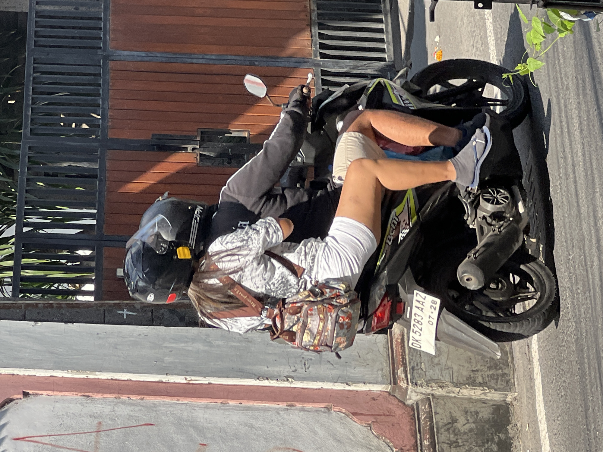 Two individuals are riding a scooter on a street in Kuta, Indonesia. The person in front, wearing a black helmet, is operating the scooter. The person behind, also wearing a black helmet, is a woman with long brown hair, carrying a patterned backpack. She is wearing a white patterned shirt and white shorts, and gray flip-flops. The scooter has a license plate that reads "DK 5283 AAZ". They are riding on a paved road, with a building wall on the left and a fence with wooden panels and greenery on the right. The lighting suggests it is daytime.
