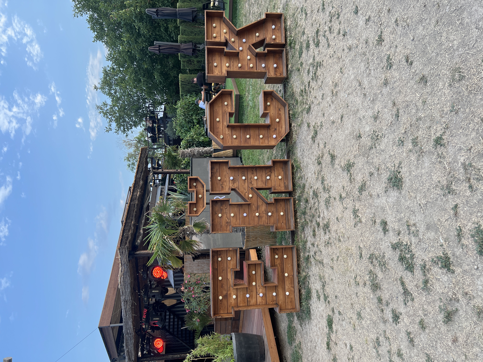 In Yeysk, Russia, large wooden letters spelling "TECK" stand on a dry, grassy ground. The letters, illuminated by round bulbs, are positioned in front of a rustic structure with a thatched roof and wooden decking. Behind the structure, palm trees and other greenery are visible, along with umbrellas and seated individuals at tables. The sky above is blue with scattered white clouds. The scene appears to be an outdoor venue, possibly a cafe or entertainment space, during daylight.