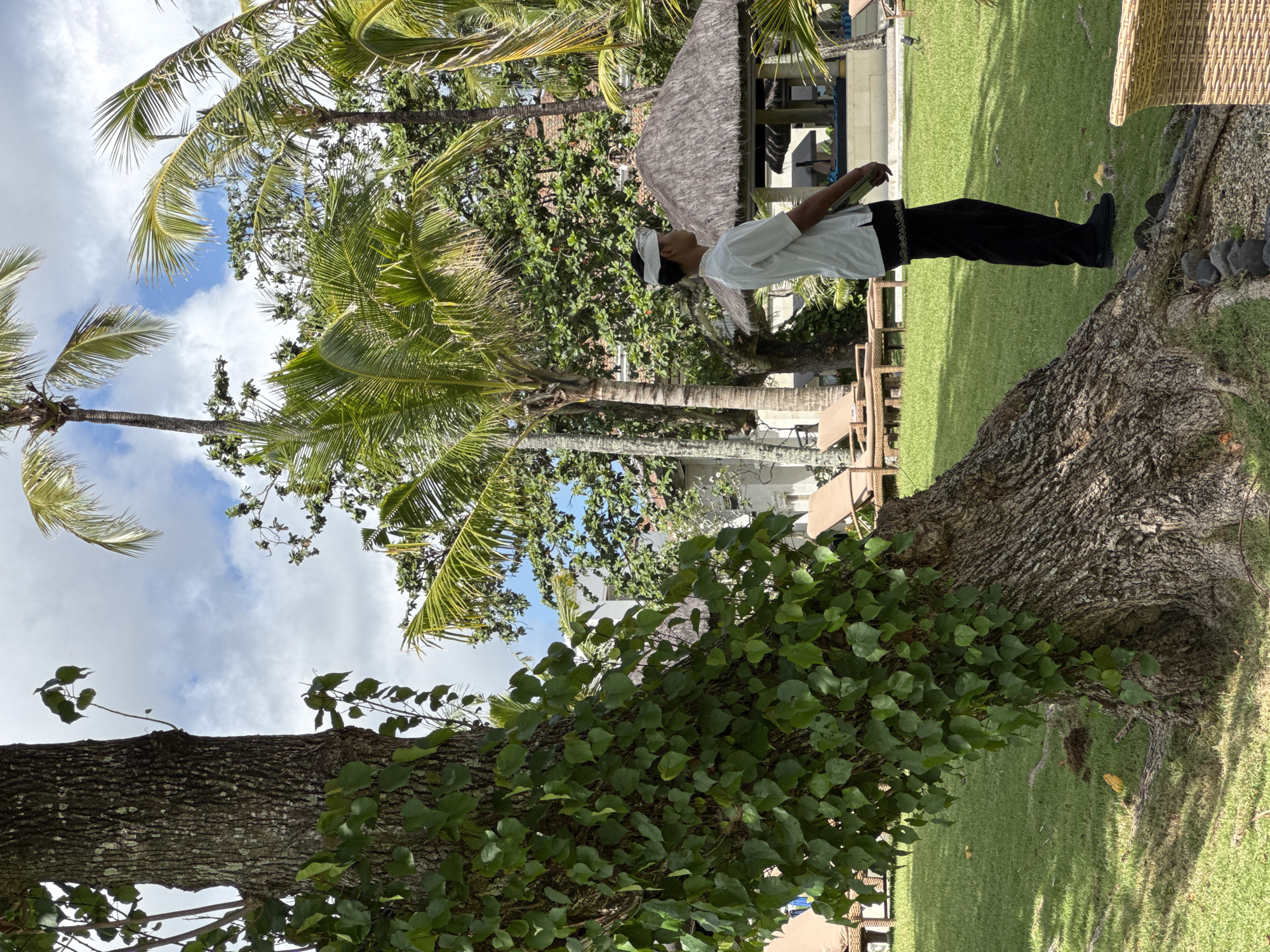 The image shows an outdoor scene in Kuta Selatan, Indonesia. A man stands on the base of a large tree, wearing a white shirt, a white headdress, and black pants with a dark patterned sash. He is holding a dark-colored object in his hands. The tree is in a grassy area, with other vegetation and palm trees in the background. The sky is partly cloudy. Some buildings and a thatched structure are also visible in the background.