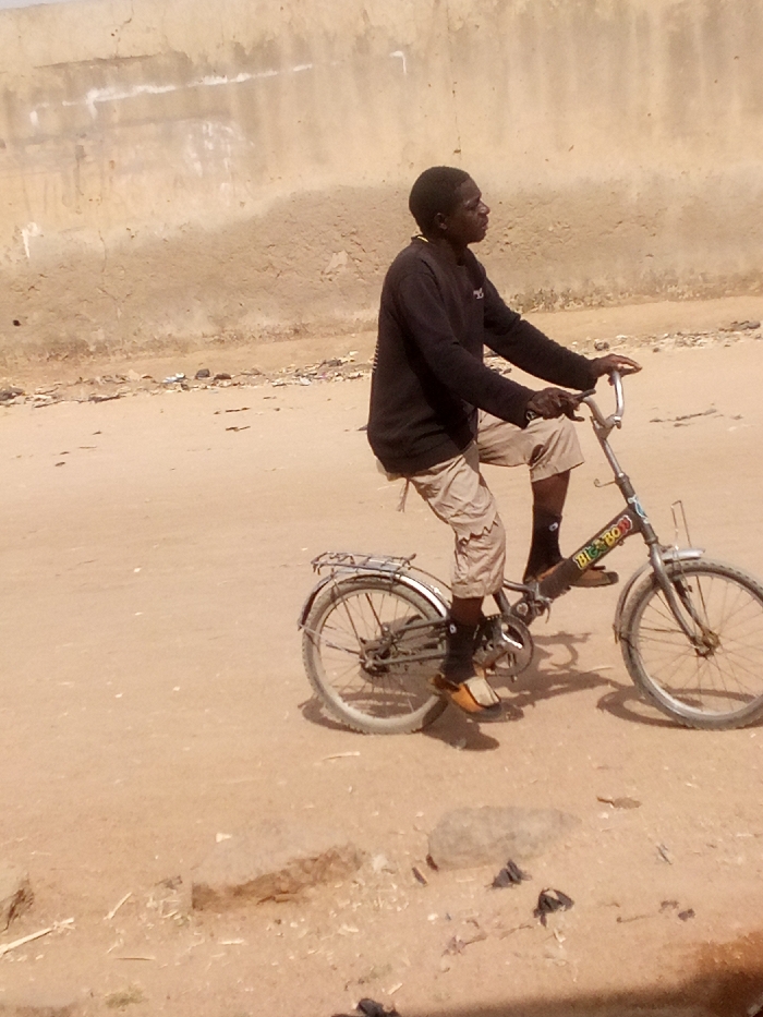 An adult male is observed riding a dark-colored "BIG BOY" brand folding bicycle on an unpaved, light brown dirt road in Tudun Murtala, Nigeria. The individual wears a dark long-sleeved shirt, light-colored knee-length cargo shorts, dark socks, and light brown closed-toe sandals. His hands are on the bicycle handlebars, and his feet are positioned near the pedals, indicating active cycling or readiness to proceed. The bicycle features front and rear fenders, a rear cargo rack, and visible wear. The road surface is uneven, strewn with small rocks and dark debris. A light brown, worn, and discolored wall or building facade forms the background, showing horizontal markings and vertical streaks. The scene is illuminated by natural daylight, casting subtle shadows beneath the bicycle and the individual.