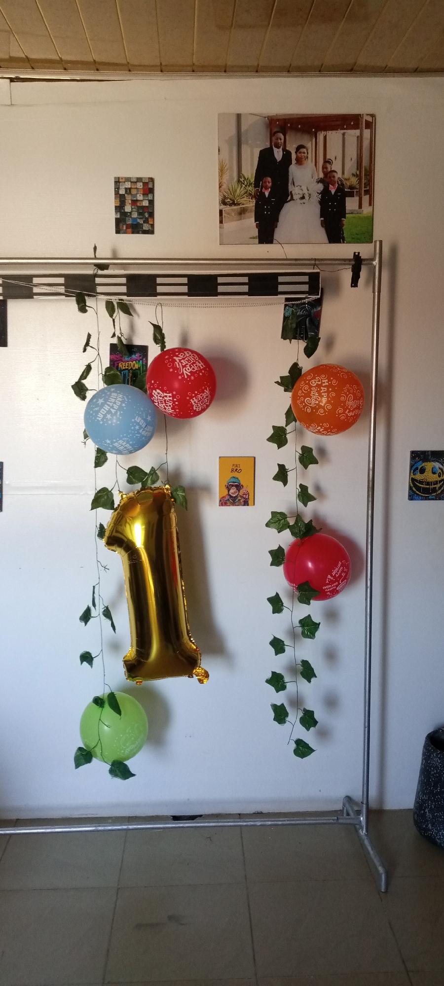 The image shows a room decorated for a first birthday, likely in a home in Jos, Nigeria, indicated by the appearance of the interior and building materials. A metal clothing rack supports decorations including: a gold foil number '1' balloon, four round balloons in blue, red, orange, and green; strands of artificial ivy; and several small printed images and cards. The background wall is white, and a photograph of a wedding or family portrait is displayed. To the left of the family portrait is a square graphic. A "peace bro" card with a monkey picture on it is visible between the balloons. A patterned floor completes the view. The scene appears to be a simple, celebratory set-up.