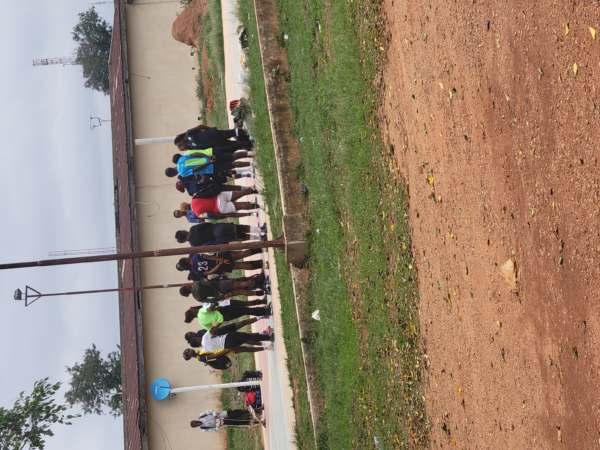 A group of people are lined up along a walkway. The location is outdoors, possibly a park or training ground. The group appears to be in some sort of formation or exercise activity. The ground surface is dirt or gravel. There are trees and buildings in the background. The location is in the city of Unknown, Unknown.