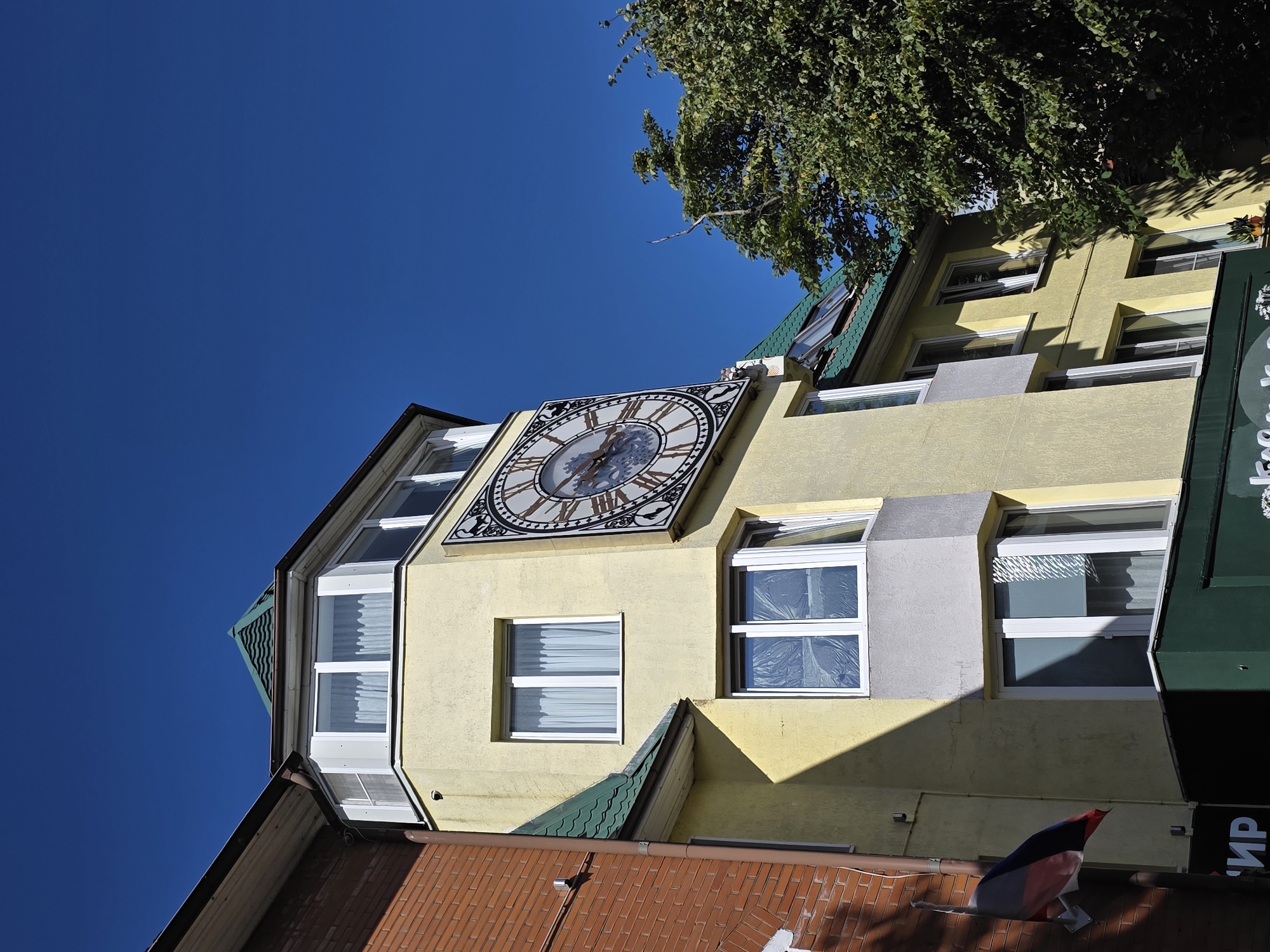 The image shows a building with a large clock on its facade, located in Zelenogradsk, Russia. The building is painted a light yellow and has a green tiled roof. Several windows are visible, some with white curtains and others covered with plastic sheeting. A Russian flag is attached to the side of the building. To the right of the building, lush green trees are in bloom. The sky is a clear, bright blue. The clock face is round with Roman numerals and ornate hands, indicating a classic design. The architecture suggests a public building, possibly a town hall or a historical landmark. The overall scene is bathed in bright sunlight, casting distinct shadows on the building.
