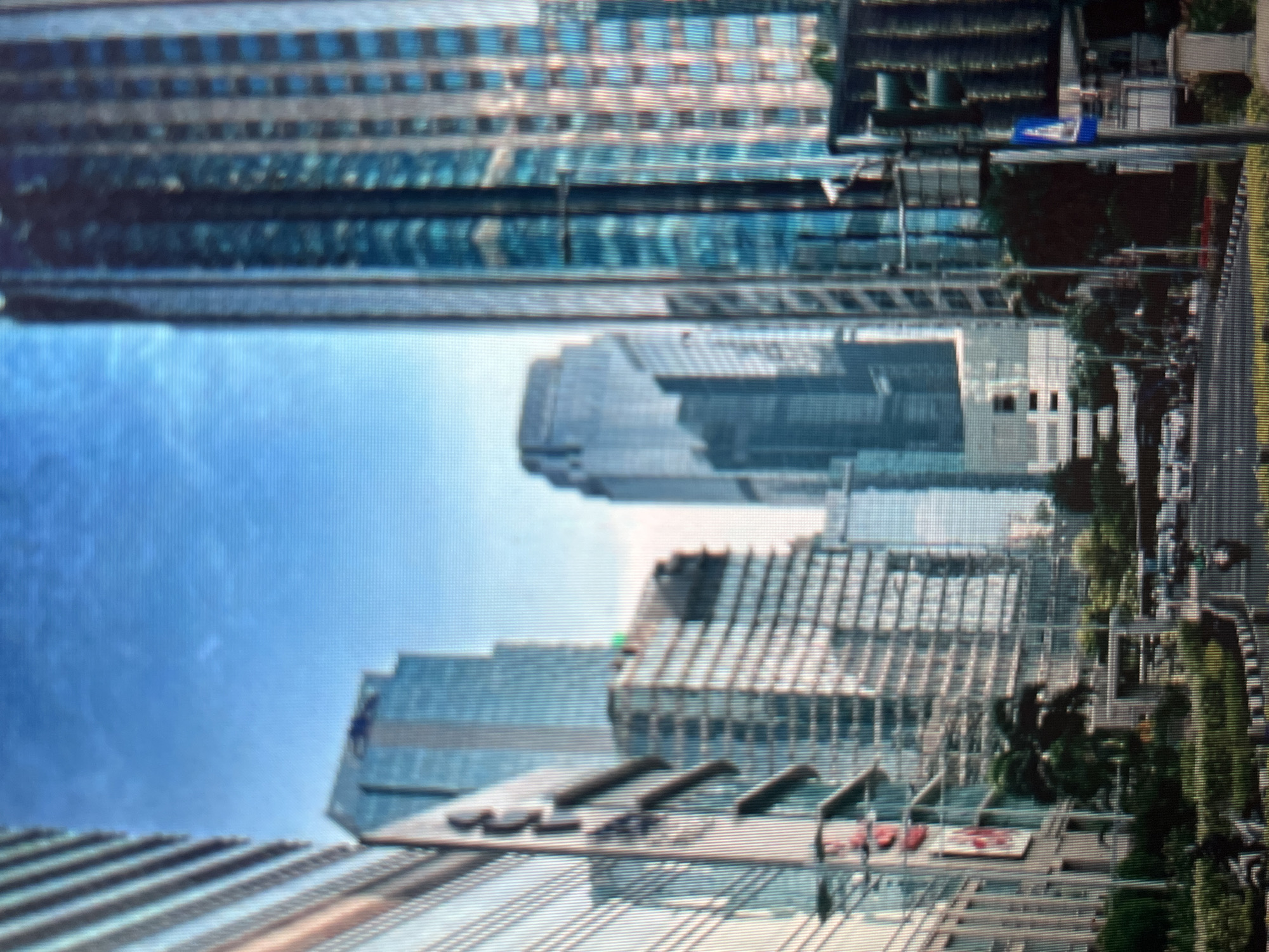 The image shows a city street scene in Sejahtera, Indonesia, with several modern glass-facade skyscrapers dominating the skyline. The buildings are tall and reflect the clear blue sky. A road with several vehicles, including cars and motorcycles, is visible in the foreground. There are traffic lights and street signs, indicating an active urban environment. Green spaces, such as trees and bushes, are interspersed among the buildings and along the roadside, adding a touch of nature to the concrete jungle. The lighting suggests it is daytime, with the sun casting shadows.